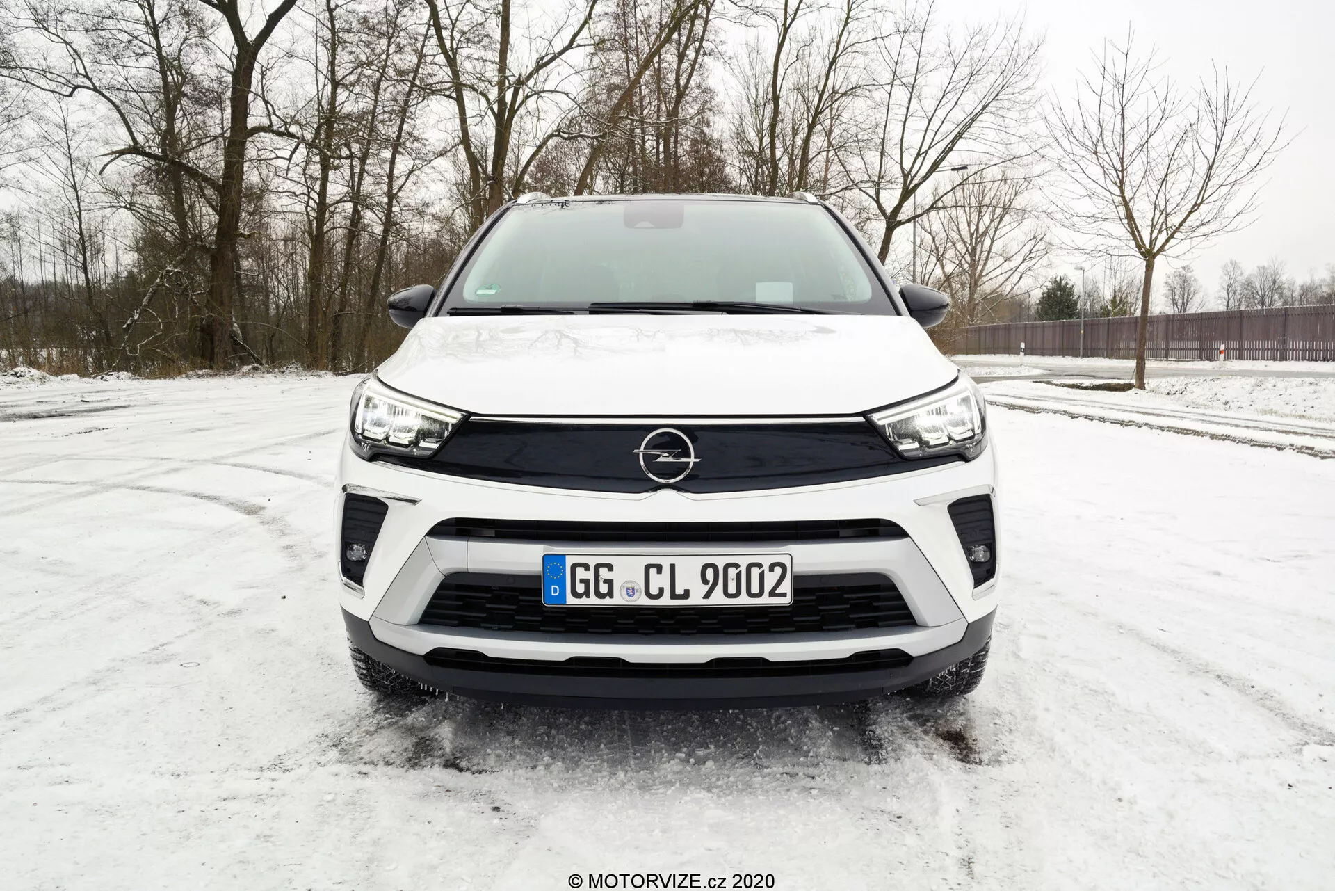 Frontal view of a modern white Opel Crossland 2021 SUV parked on a snowy surface, with a low-angle shot highlighting the front grille, headlight design, and black bumper details. Key features include the prominently displayed Opel emblem, sleek LED headlights, standard license plate placement, potential integration of parking sensors, and a robust bumper design suggesting off-road capability.