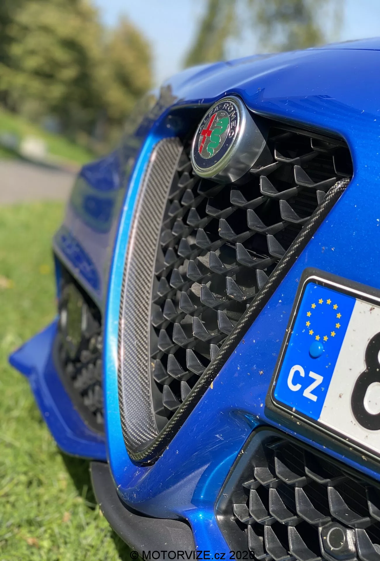 Close-up view of Alfa Romeo Giulia 2016's front end from a low angle, highlighting the honeycomb mesh grille, central emblem, aerodynamic front bumper, and glossy metallic blue bodywork with reflections. Partial view of a Czech license plate and blurred park-like background.