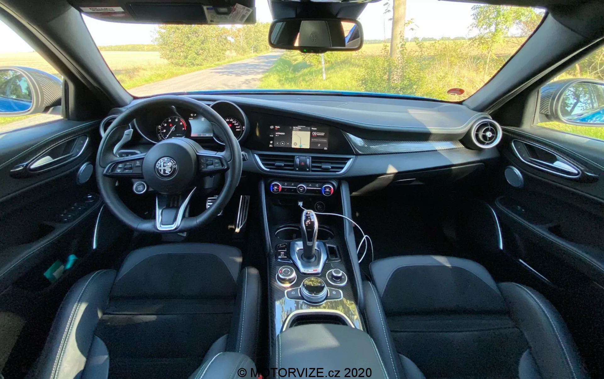 Interior view of a 2016 Alfa Romeo Giulia from a driver's perspective showcasing a flat-bottom steering wheel with multiple control buttons, a digital infotainment system on the dashboard, an analog clock, circular air vents, a sleek gear shift lever, driving mode selector, cup holders, electrically operated windows, leather seats with side bolstering, and overall modern technology features.