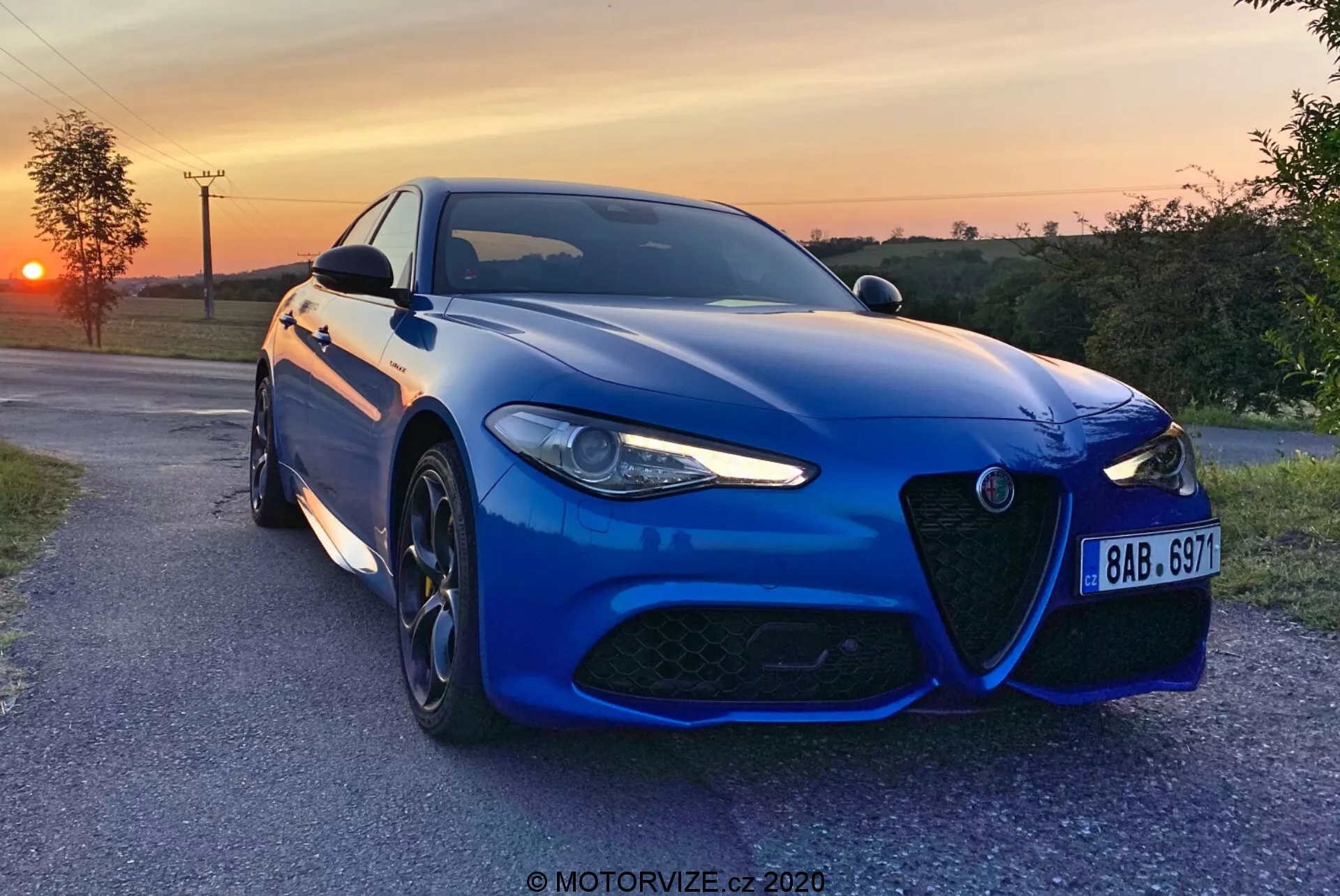 A blue Alfa-Romeo Giulia 2016 sports sedan captured in the evening with a sunset in the background, positioned slightly angled towards the camera on a paved countryside lane, showcasing its distinctive front grille, aggressive LED headlight design, large multi-spoke alloy wheels, and high-performance braking system. The metallic paint, aerodynamic front bumper, sculpted hood lines, and advanced driving assistance sensors highlight the car's sporty aesthetics and modern technology. A watermark 'MOTORVIZE.cz 2020' is present.