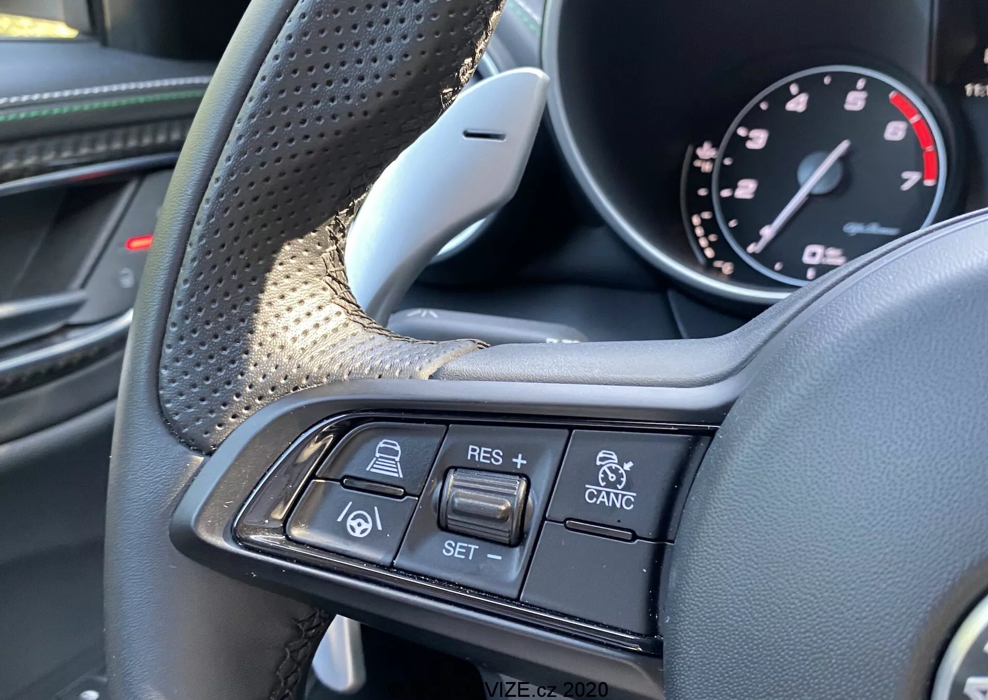 Close-up view of the Alfa-Romeo Giulia 2016 interior showcasing the steering wheel and its multifunction controls from the driver’s perspective. The textured grip areas and luxurious materials like leather and possible carbon fiber are visible on the left side. The right spoke features cruise control and adaptive cruise technology buttons, including + RES, - SET, CANCEL, a car-following-car icon, and vehicle collision alerts. The image focuses exclusively on these controls, emphasizing the premium design and advanced driver assistance features.