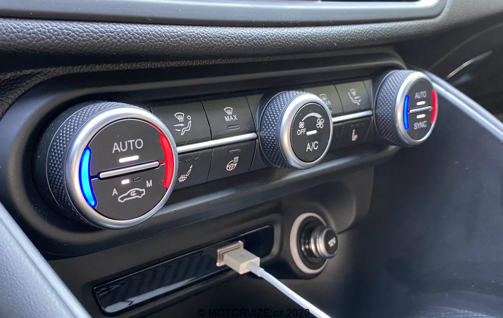 Close-up of the climate control system on the dashboard of a 2016 Alfa Romeo Giulia, featuring three circular knobs. The left knob indicates temperature settings with blue and red bands and an 'AUTO' setting, partially lit in red. The middle knob shows fan speed settings, highlighted at a lower speed. The right knob displays various air distribution modes with 'AC' and 'SYNC' buttons. Below the knobs are a 12-volt power outlet and USB port. The dashboard has a black finish with a silver accent around the climate controls, captured from an elevated angle.