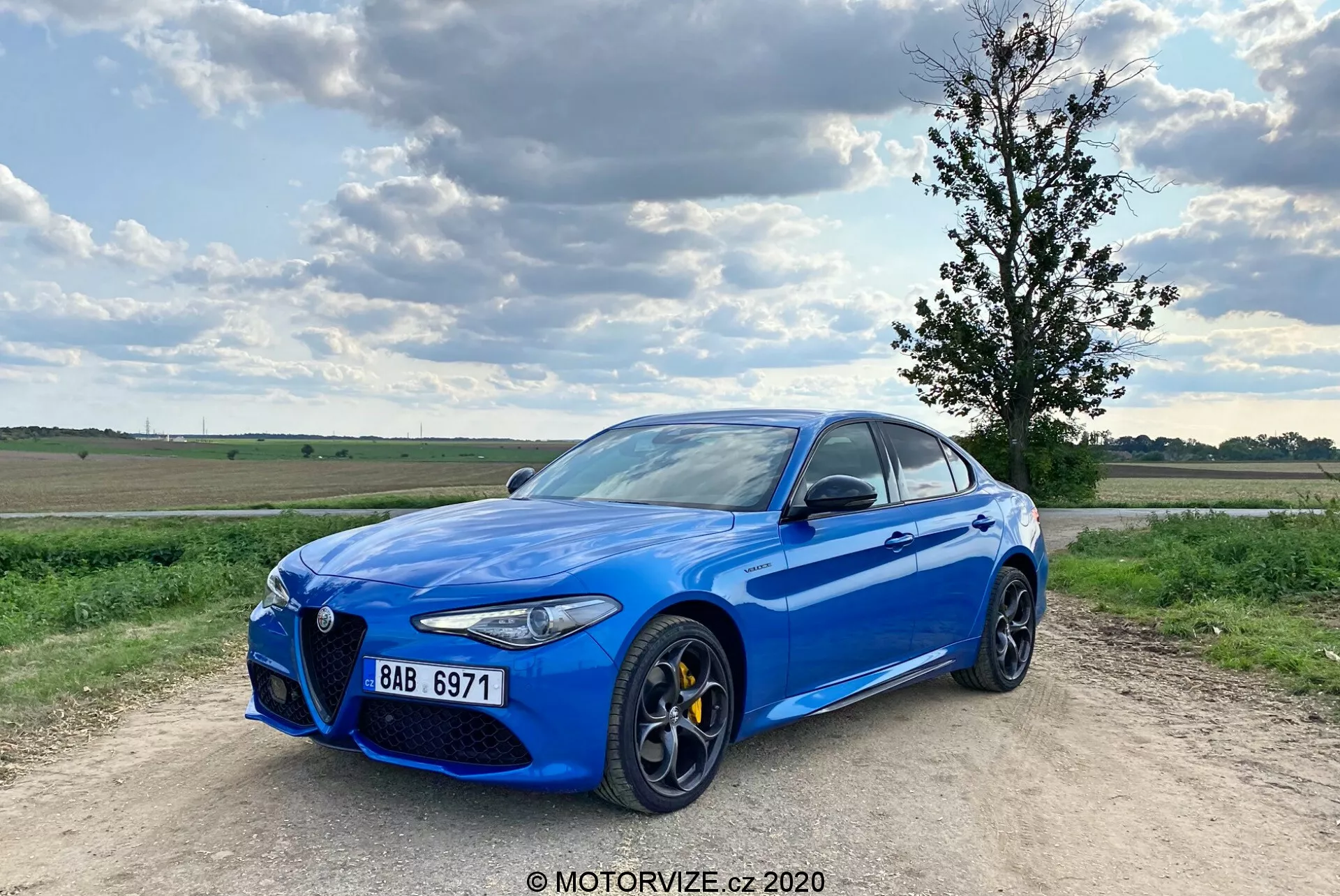 Image of a bright blue Alfa Romeo Giulia 2016 sports sedan parked on a gravel path, with a partially cloudy blue sky in the background. The car is captured from a three-quarter front angle, showcasing its distinctive 'Trilobo' front grille, yellow brake calipers, large multi-spoke alloy wheels, sleek LED headlamp clusters, and chrome window surrounds. The vehicle is slightly off-center to the left with the front left wheel pointing outward, giving it a dynamic stance.