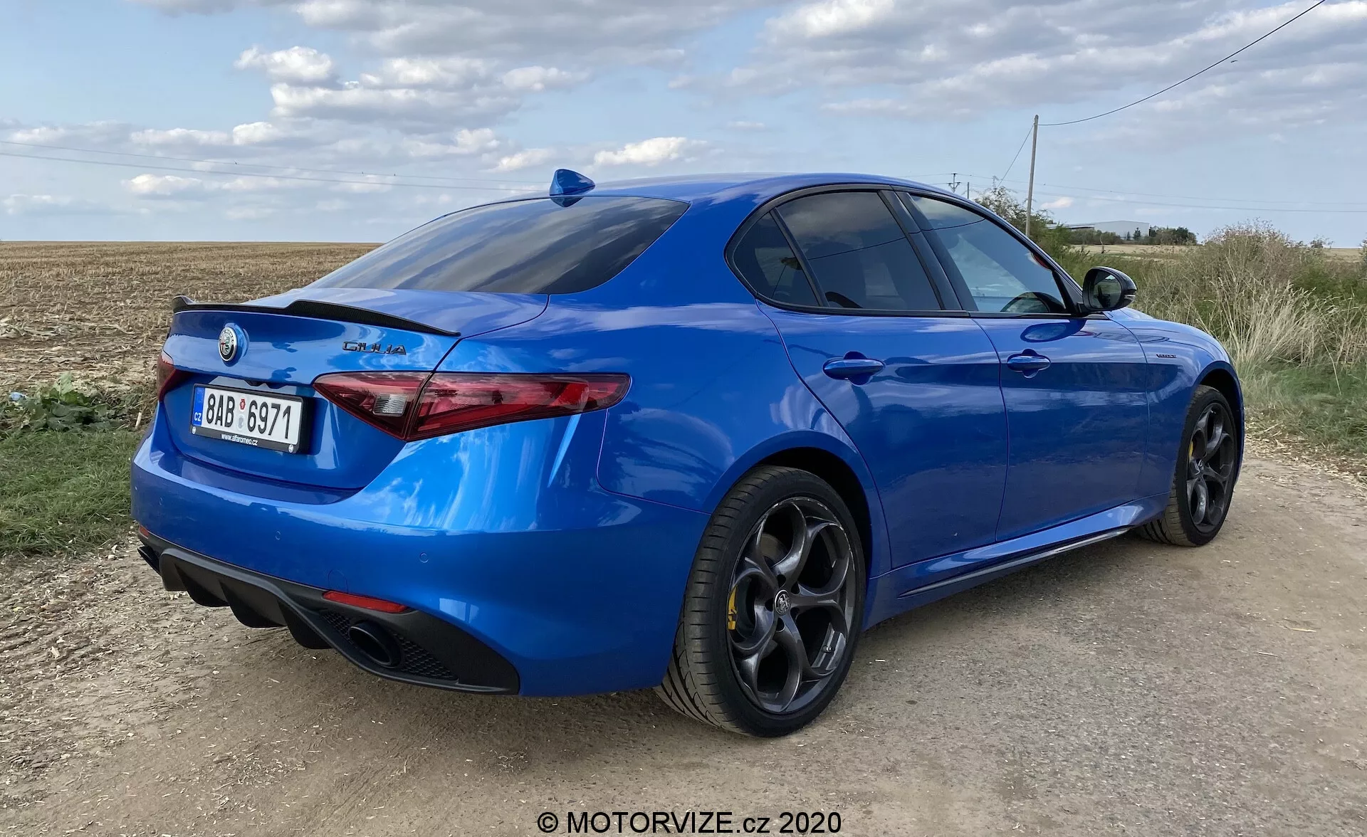 Rear three-quarter view of a bright blue sports sedan on an unpaved road with open field and partly cloudy sky in the background. Low-angle photograph highlighting the car's stance, sculpted rear bumper with integrated dual exhausts, slim horizontally oriented rear light clusters, lip spoiler on the trunk lid, and black multi-spoke alloy wheels with low-profile performance tires. The side profile shows a sleek silhouette with a sloping roofline and slightly tinted rear windows. The rear of the car features a badge and brand emblem, indicating model or trim level. The car's exterior appears in excellent condition, and the ample daylight provides clear visibility of the car's design and paintwork.