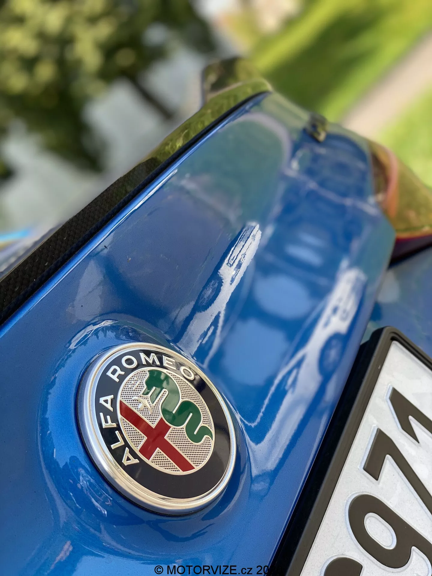 Close-up view of Alfa Romeo Giulia 2016 rear section featuring brand emblem from low angle. The badge, with a red cross on white background and green snake on blue background, is in sharp focus on metallic blue boot lid, with surrounding surface details and reflections.
