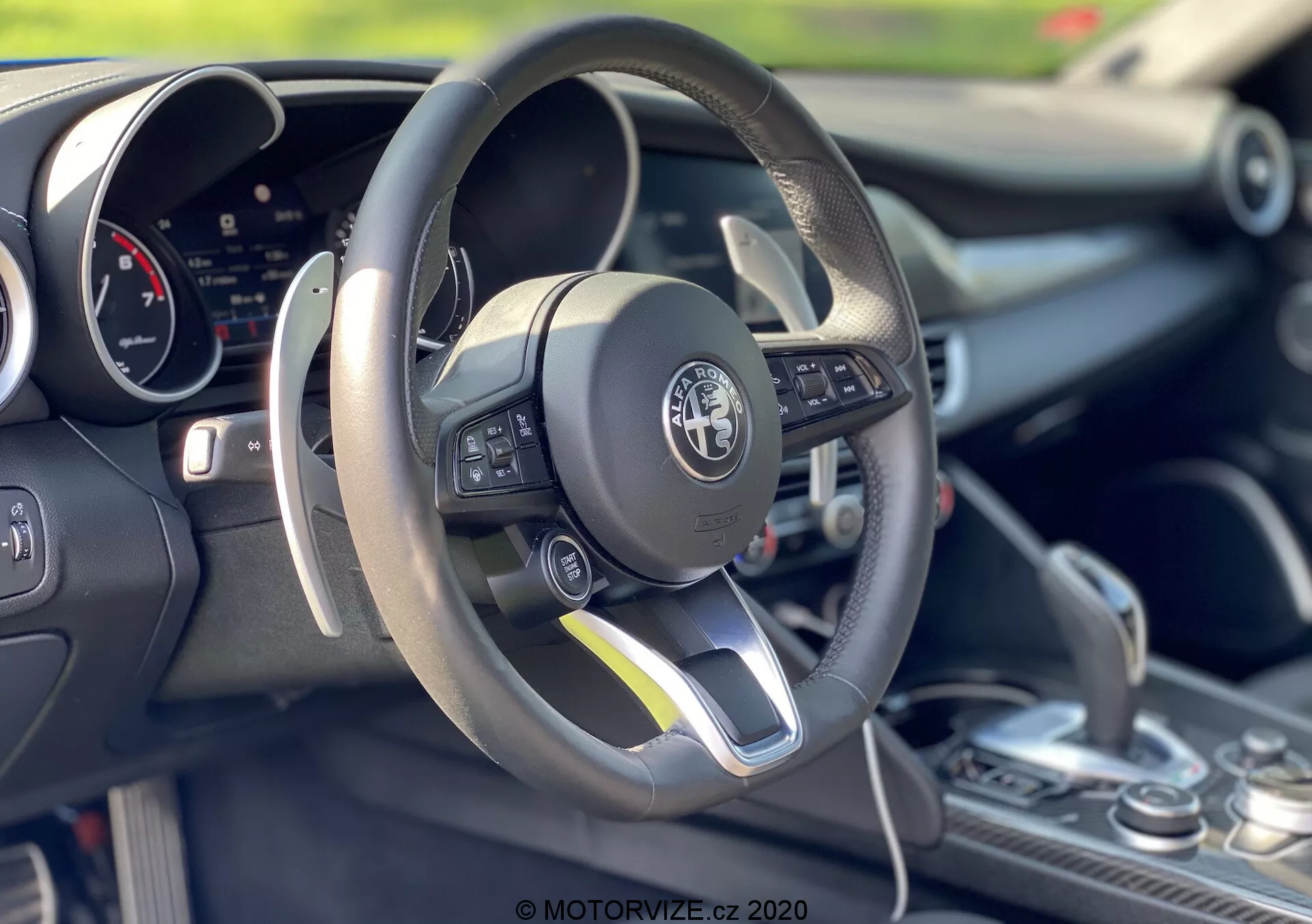 Interior shot of a 2016 Alfa-Romeo Giulia focusing on the driver's area and steering wheel. The flat-bottom, leather-wrapped steering wheel features multifunction controls on the left-hand side. A digital instrument cluster is visible behind the wheel. The center console shows a gear shift with a leather boot, suggesting possible manual shift capabilities. The image emphasizes the car's sporty design, advanced technology, and detailed craftsmanship with a shallow depth of field highlighting the forefront elements.