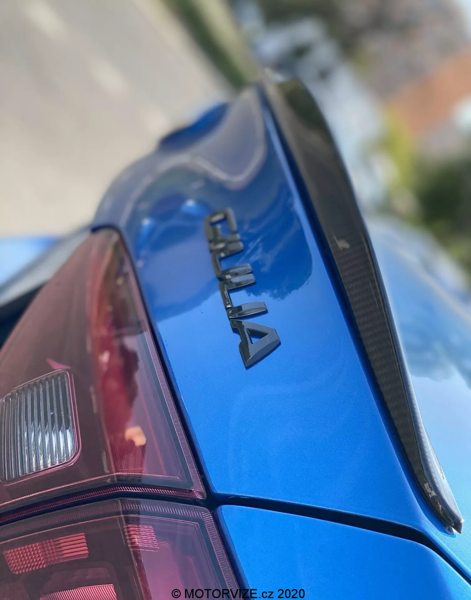 Close-up image of the rear section of a blue ŠKODA car, viewed from an upward angle. The focus is on the chrome 'ŠKODA' badging on the trunk lid, a slim spoiler, and the glossy, well-maintained paintwork. The rear windshield features defroster lines, and part of the modern-designed right rear light cluster is visible. The shallow depth of field emphasizes the branding, with the front and background blurred. Watermarked 'MOTORVIZE.cz 2020', indicating its publication or capture date.
