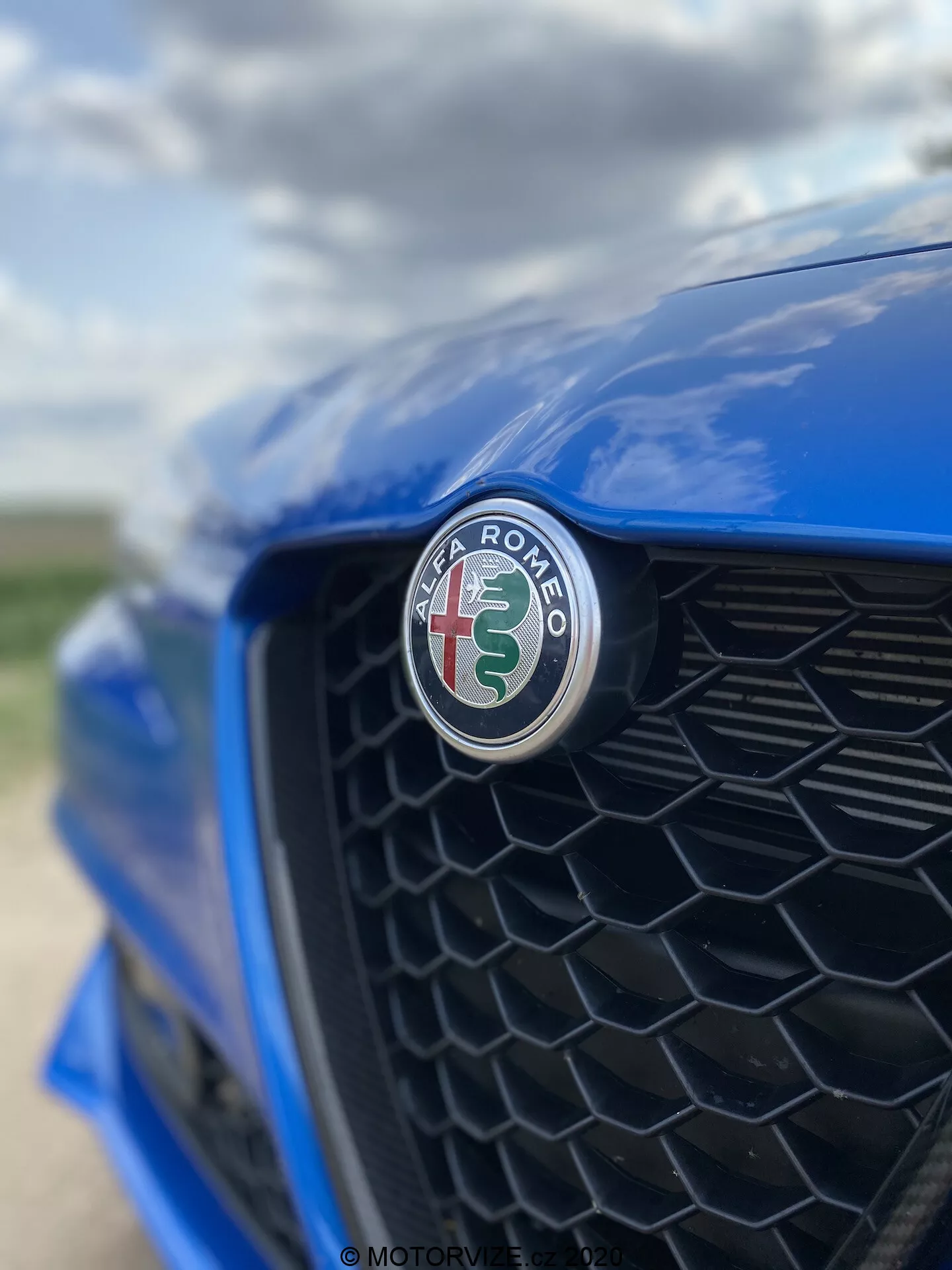 Close-up of the front section of a blue Alfa Romeo Giulia 2016, focusing on the manufacturer's emblem featuring a red cross and green serpent over a white background. Shot from an off-center angle towards the driver's side, the black honeycomb grille is softly defocused, highlighting the emblem's sharp focus and glossy finish.