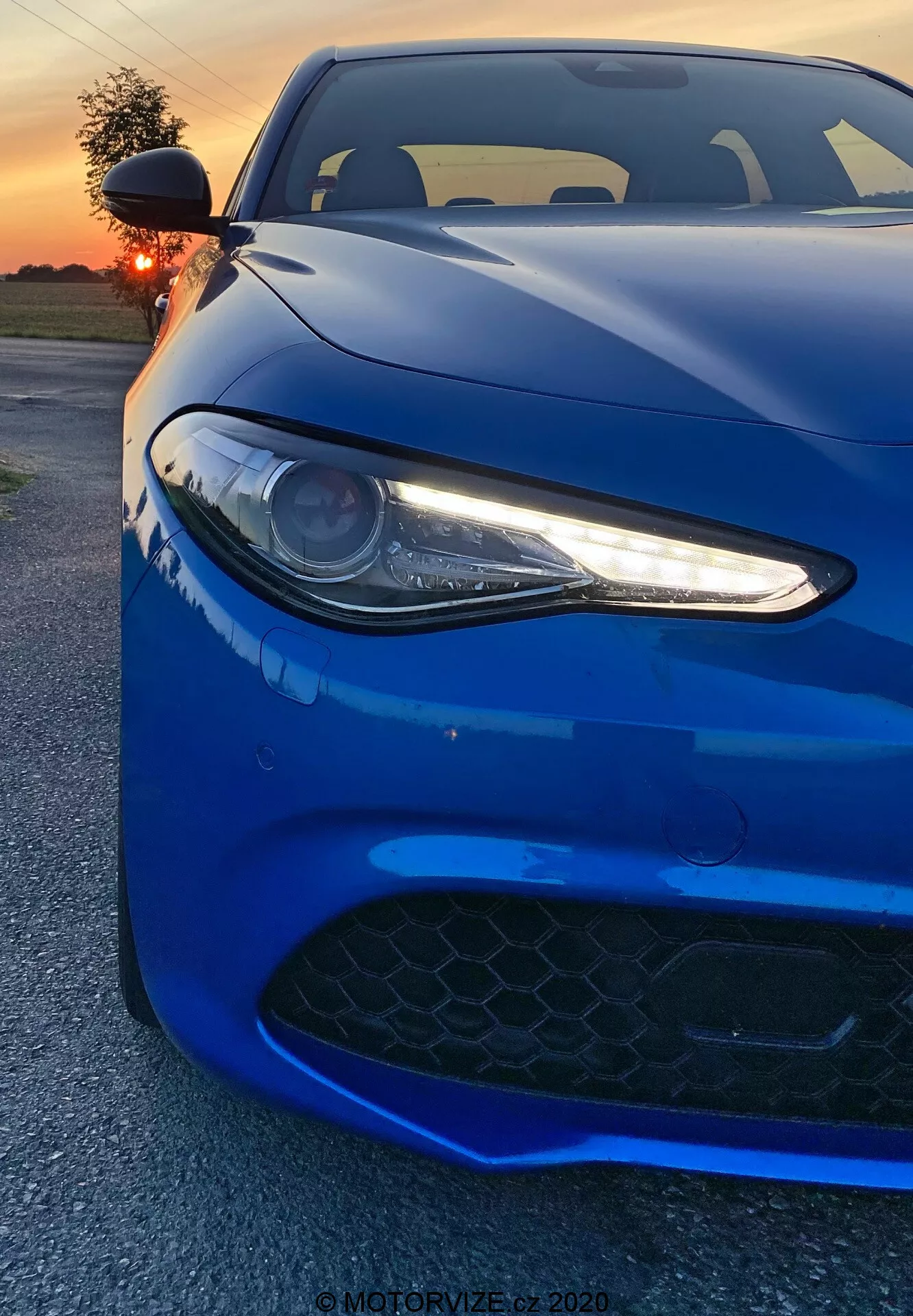 The image depicts a 2016 Alfa Romeo Giulia sports car captured from a low front angle. The vibrant, deep blue vehicle fills the frame, emphasizing its left headlamp with LED daytime running lights, a part of the hood, and the air intake grille in the front bumper. The projector-type headlight, with its sleek white LED accents, gives an aggressive, modern look. The black hexagonal-patterned grille suggests high-performance capabilities. The shot captures the car's aerodynamic lines and contours, with reflections from a sunset enhancing its glossy finish. The background shows a warm, orange-lit sky partially obscured by the sun. The photographer's close angle showcases design details, evoking a sense of power and elegance.