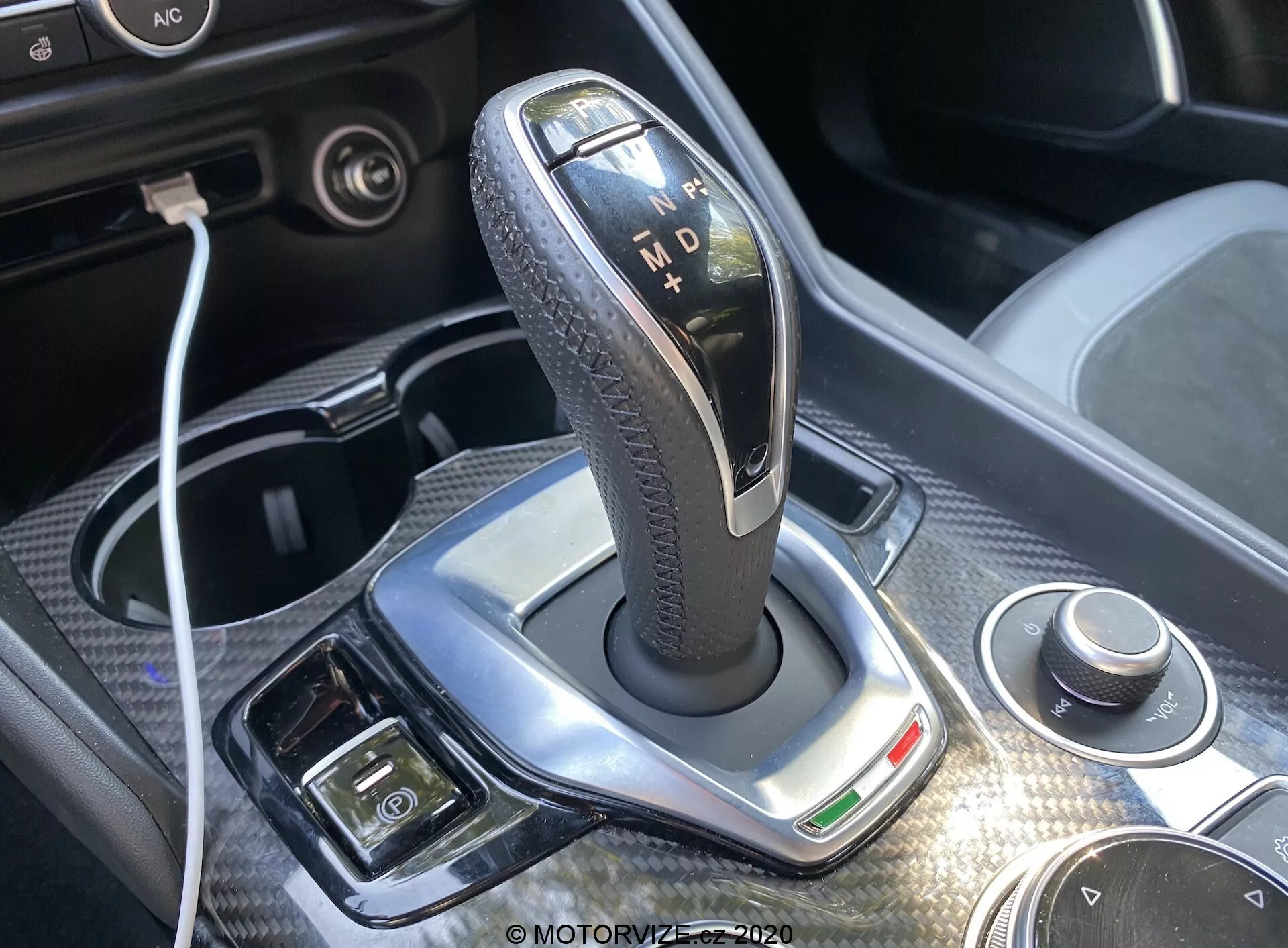 Close-up of the gear shift lever in a 2016 Alfa-Romeo Giulia's automatic transmission, featuring a sleek, polished silver surround, leather-textured black grip, and illuminated indicators for 'P', 'R', 'N', 'D', and 'M'. Bright daylight enhances the design, showcasing a rotary dial and USB connectivity nearby. Chrome-like finish, green indicator light, and additional buttons for electronic parking brake and climate control are visible, offering a glimpse into the luxury and advanced technology of the car's interior.