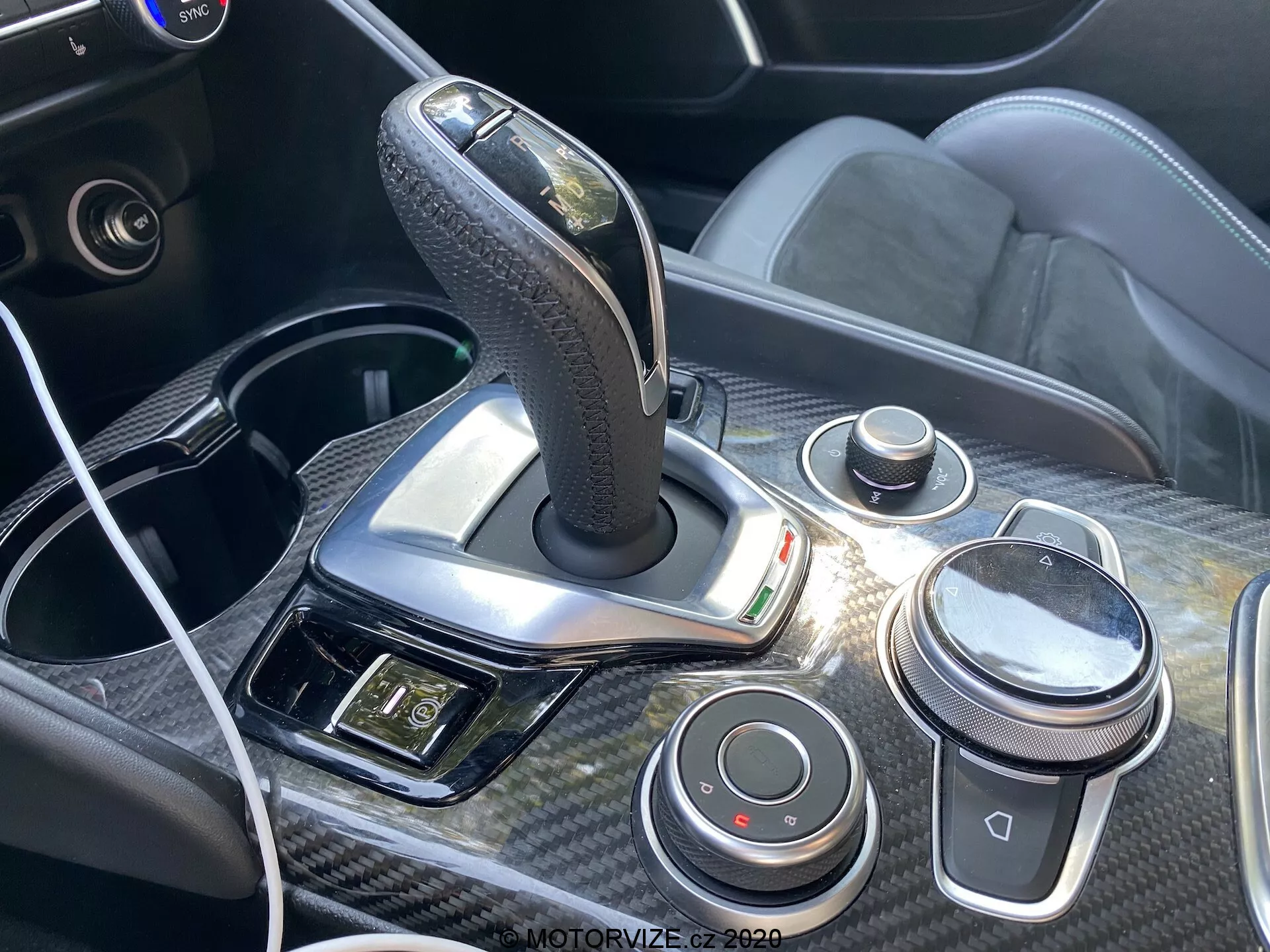 Close-up view of the 2016 Alfa Romeo Giulia's center console and gearshift area featuring a leather-wrapped automatic gearshift with chrome accents, surrounded by carbon-fiber trim. The console includes iDrive-like control knob, volume control dial, driving dynamics buttons, USB port with a connected cable, and a panel with hazard light and other controls, emphasizing the car's ergonomic and high-tech interior design.