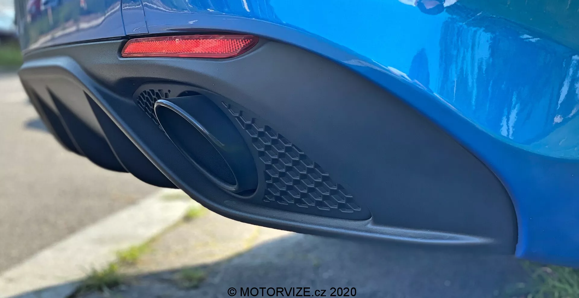 Close-up side view of the rear bumper and exhaust area of a vibrant blue Alfa Romeo Giulia 2016, showcasing its modern design features including an elongated horizontal red LED tail light, large oval-shaped exhaust port with patterned mesh detail, and aerodynamically styled bumper with various curves and indentations, photographed with a shallow depth of field to emphasize the car's details.