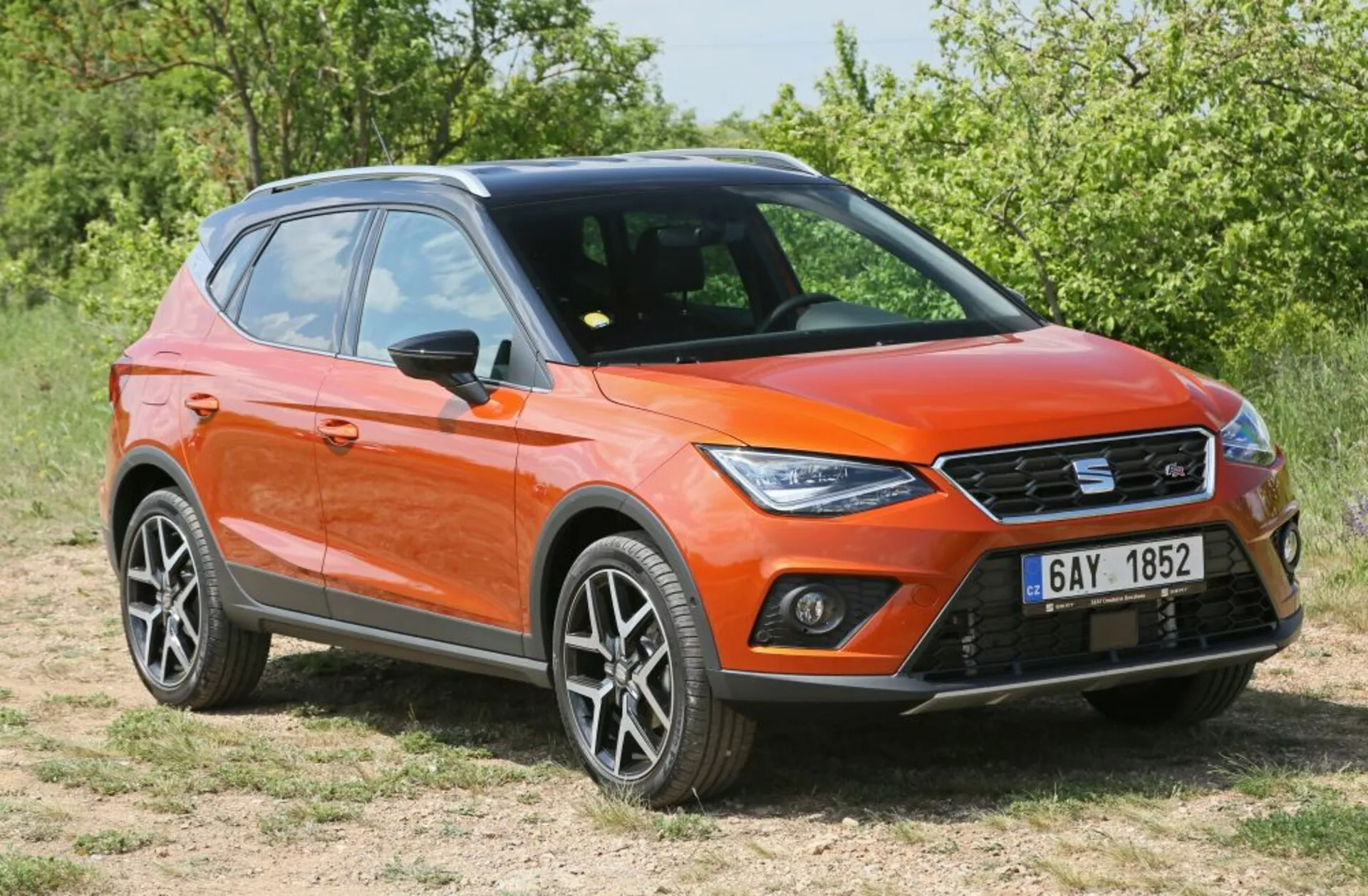Orange crossover/SUV parked on grass in front three-quarter view displaying LED daytime running lights, modern sharp design with alloy wheels, and roof rails under sunlight