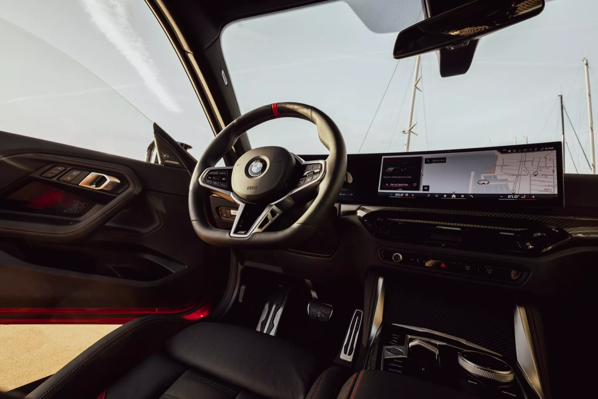 Interior view of the BMW 2er-reihe 2024 (G42-G87 Facelift) from the passenger seat. The image shows a three-spoke steering wheel with integrated controls, a digital instrument cluster, a large infotainment touchscreen, and minimalistic climate controls. Ambient red lighting is visible, and the automatic transmission selector is located in the central console. The door panel features window and mirror controls along with a speaker grille. Leather seats with contrasting stitching add to the luxury feel.