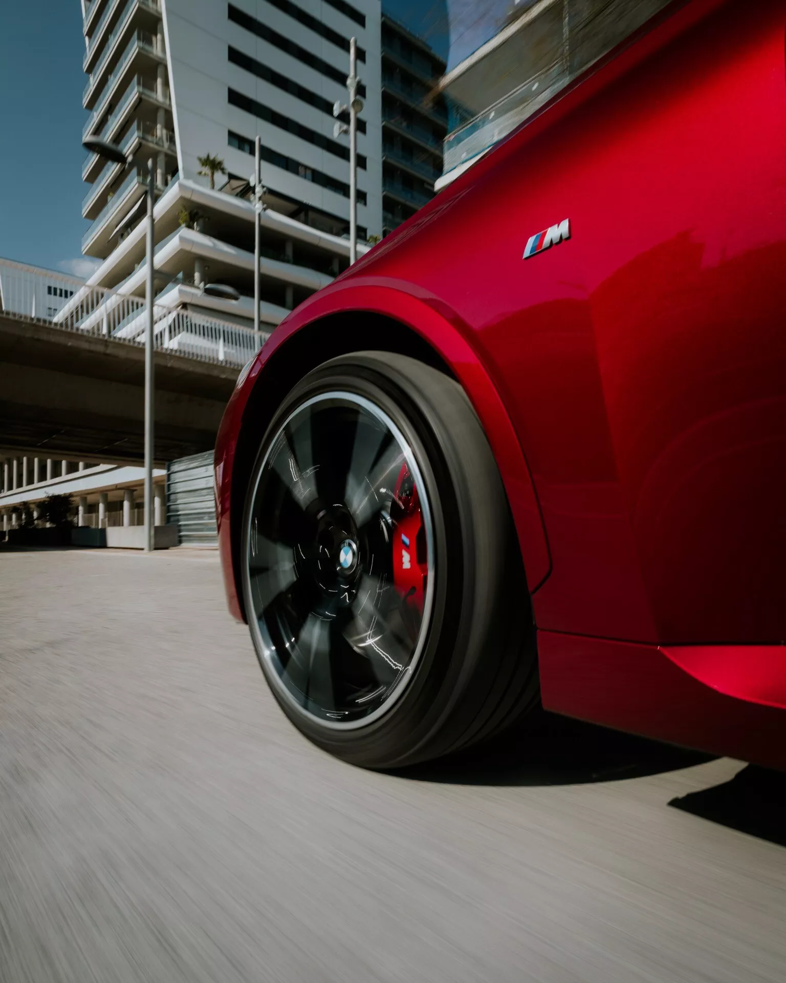 Close-up of the rear quarter of a vibrant red 2024 BMW 2 Series (G42-G87 facelift), showcasing the dynamic view from a low angle with a focus on the rear black alloy wheel featuring 5 double spokes and a contrasting red rim edge line. Prominent red 'M' badging indicates high-performance, sporty design. The spinning wheel and blurred urban background emphasize motion and speed, with drilled brake rotors and red calipers hinting at high-performance braking.