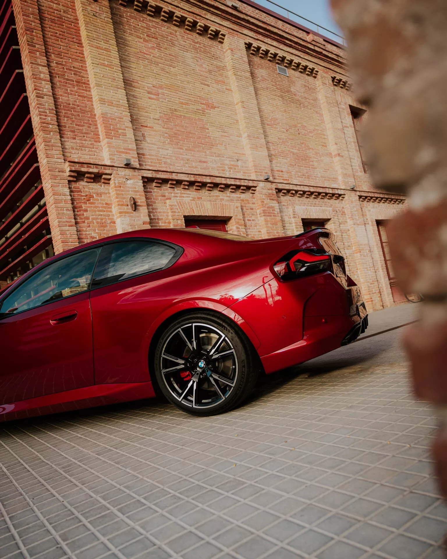 2024 BMW 2 Series (G42-G87 Facelift) in red, captured from a low side angle, showcasing the car's full profile including front wheel, door, side vent, and rear wheel. The image highlights high-performance wheels, aerodynamic side mirrors, sleek coupe body style, large brake calipers, and rear spoiler. Photographed with a shallow depth of field, the blurred background features a historical building, emphasizing the car as the main focus and highlighting its design lines, proportions, and reflections on the bodywork.