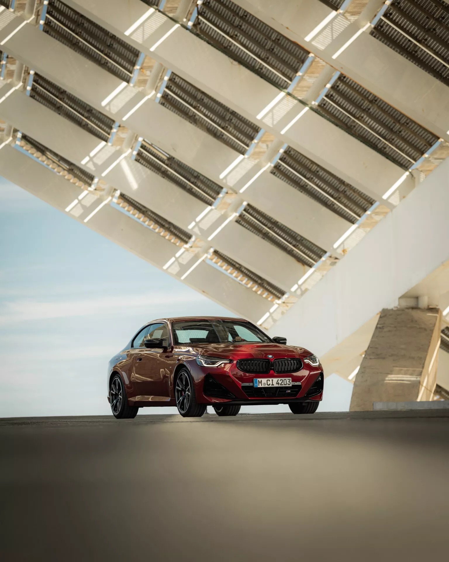 The image shows a 2024 BMW 2 Series (G42-G87 facelift) luxury sports sedan from a low angle, highlighting its stature and design lines. Captured from the front three-quarters view, it displays the car's driver's side, showcasing the front grille, LED headlights, contour of the hood, side profile, wheels, and doors. The sedan is positioned on a sleek paved area with a modern geometric-architectural structure overhead, likely part of a bridge or elevated highway, featuring metallic elements that emphasize the car's sleek design and color. The aerodynamic silhouette, large diameter sporty rims, and streamlined exterior mirrors hint at the vehicle's advanced aerodynamics and modern technology.