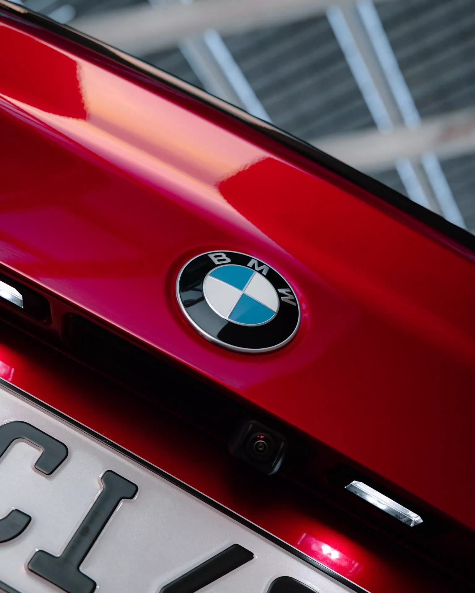 Close-up view of the rear portion of a shiny red 2024 BMW 2 Series (G42-G87 facelift), showcasing the BMW badge centrally located on the trunk lid. Below the emblem, a partially visible license plate and a small rear-facing camera are present, highlighting technology features. Two small red lights under the badge could be additional brake lights or rear fog lights. The photo's upward angle and tight framing emphasize the brand identity and sleek design elements, with a bokeh effect background.