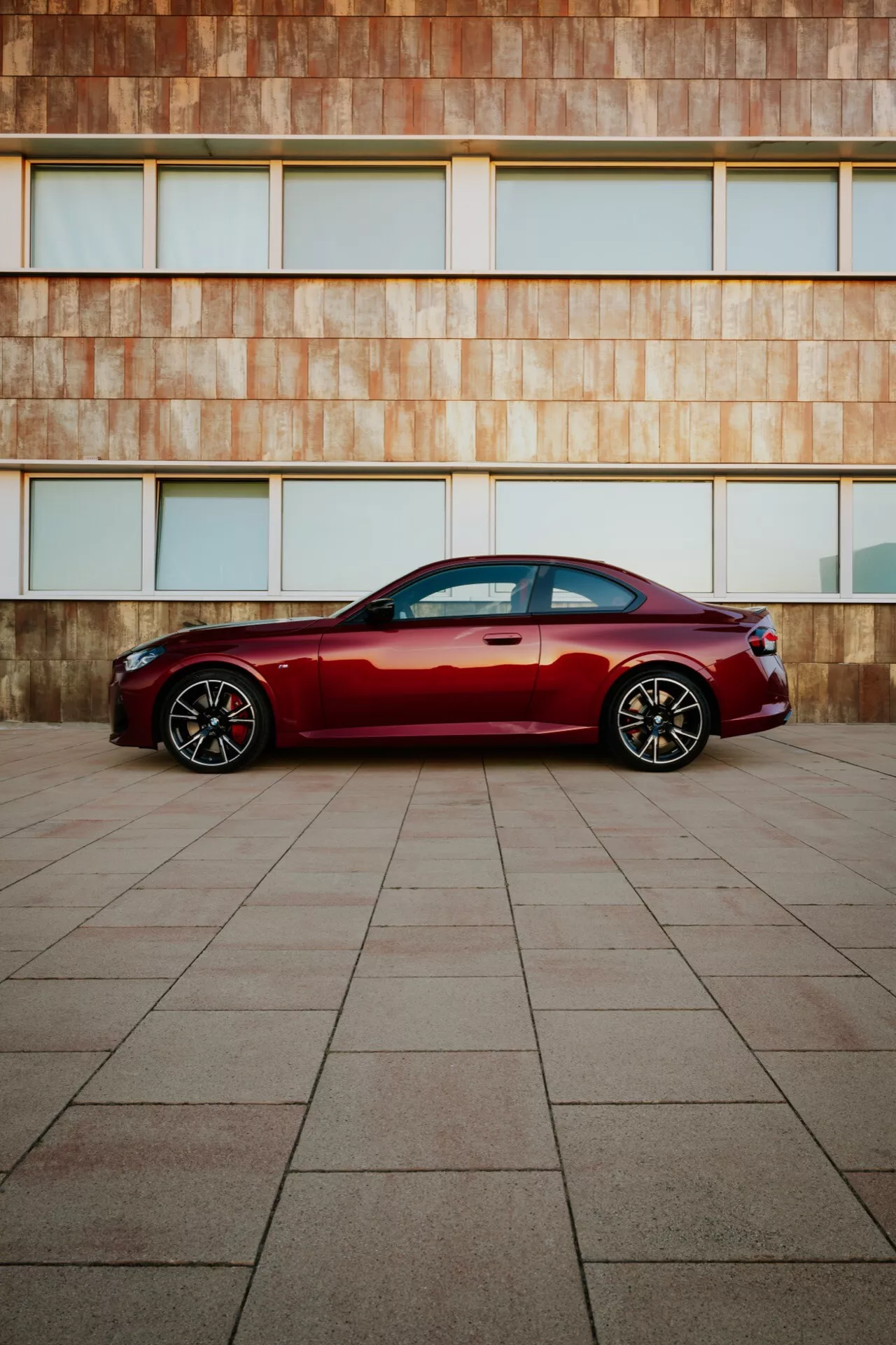 Modern luxury sedan deep red BMW 2er-Reihe 2024 (G42-G87 Facelift) parked on large tiled surface with rusty hue architectural building facade background, side profile view, sleek lines, chrome window trim, sporty alloy wheels, natural lighting.