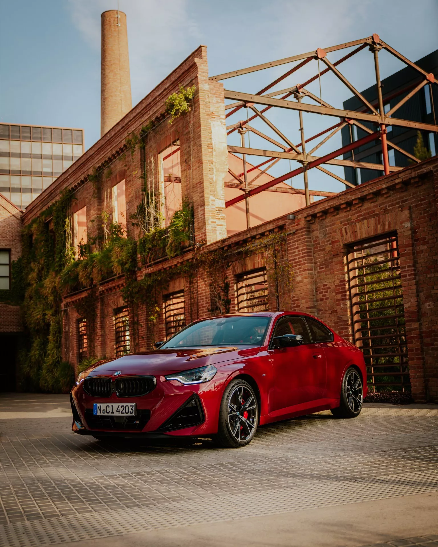 A low-angle shot of a red 2024 BMW 2 Series (G42-G87-facelift) parked in an industrial or warehouse district, showcasing its sporty front bumper with large air intakes, sleek narrow headlights, kidney-shaped grille, and aerodynamic side mirrors. The image also features multi-spoke black alloy wheels with low-profile tires and visible brake calipers. The sculpted hood lines and glossy paint finish are highlighted by warm ambient light, emphasizing the car's luxurious and performance-oriented design elements.