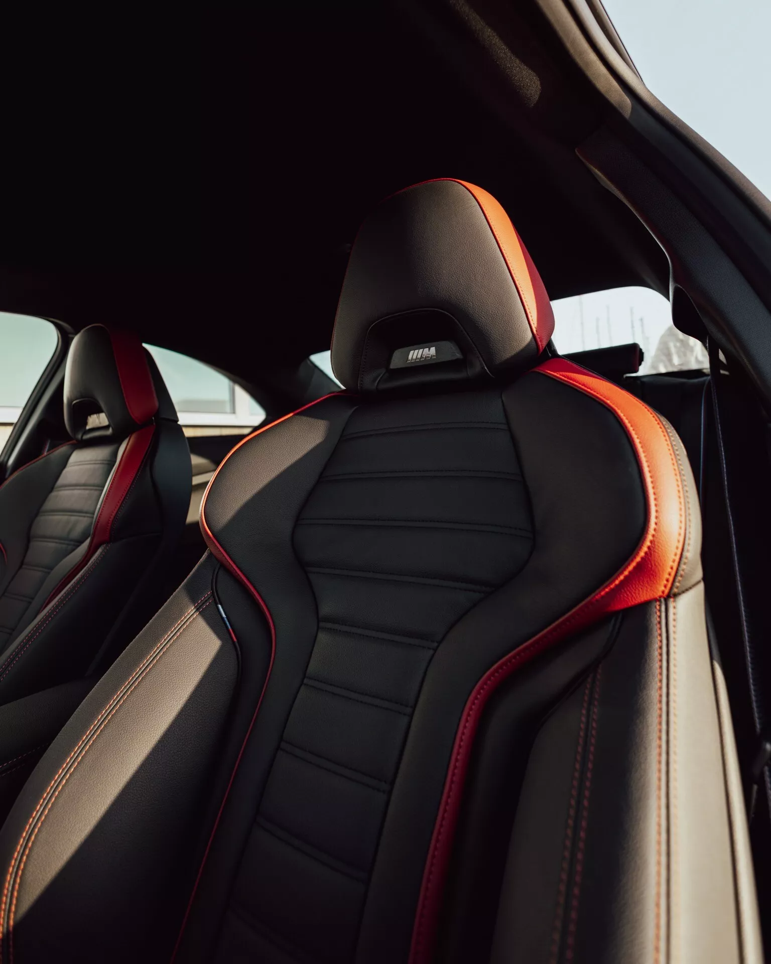 Close-up view of the BMW 2er-Reihe 2024 (G42-G87 Facelift) front passenger seat from the rear passenger compartment, showcasing the luxurious black leather upholstery with red stitching and piping. The ergonomic design with prominent side bolsters, a headrest embroidered with a performance or luxury sub-brand logo, all set against natural daylight.