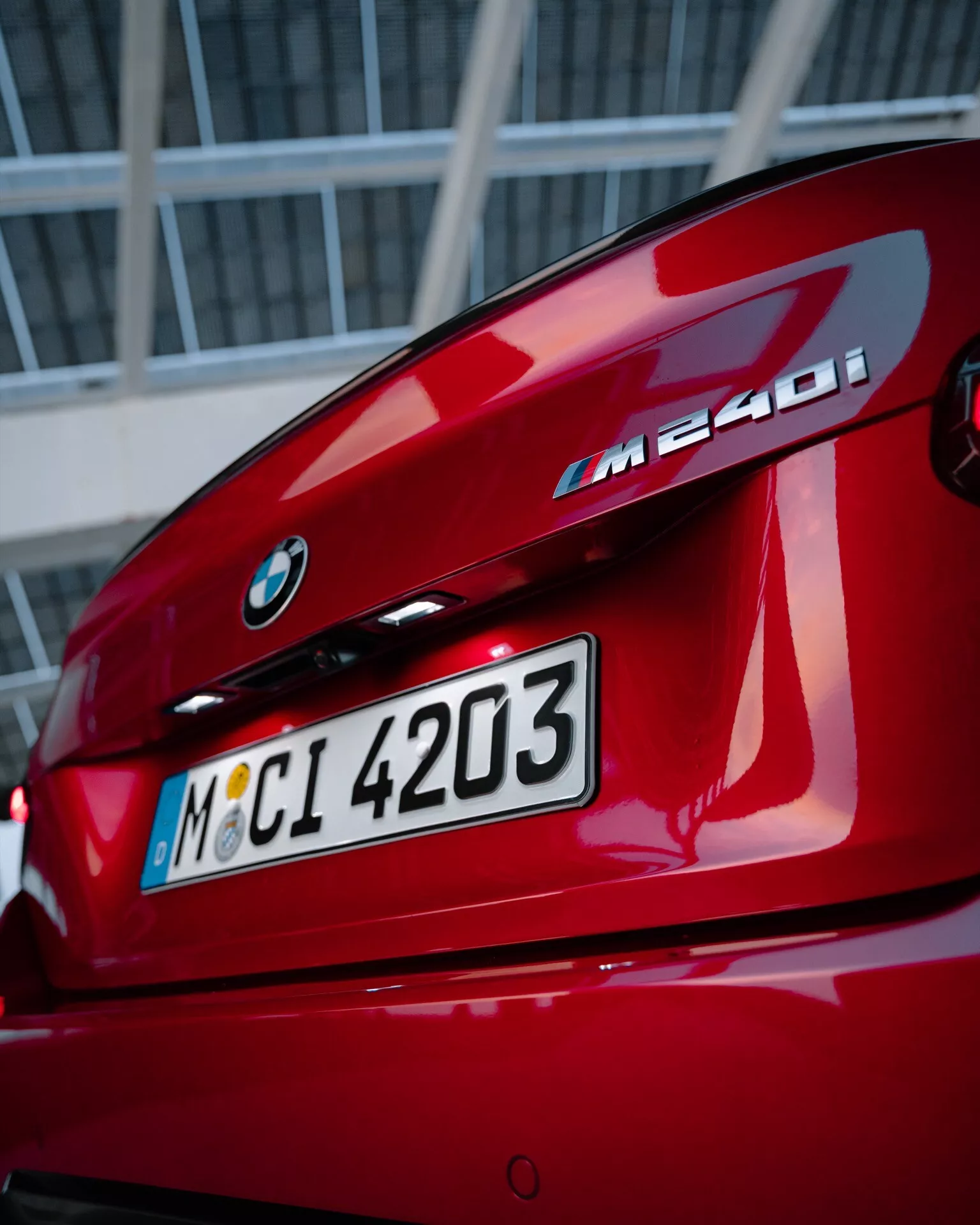 Close-up view of a red BMW M240i rear, showcasing BMW emblem, model designation, trunk lid, L-shaped taillights and integrated rear spoiler, with urban or industrial backdrop.