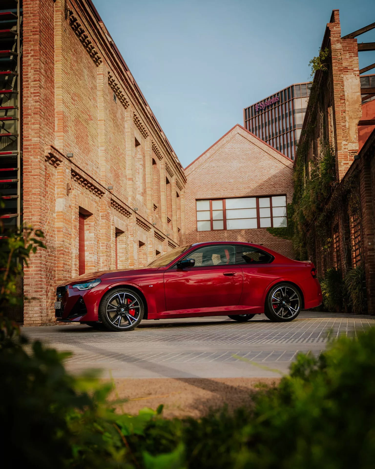 A red BMW 2 Series 2024 (G42-G87 facelift) parked in an urban setting, with brick buildings in the background. The car is shown from a profile view with design lines across the doors and fenders, and sporty alloy wheels clearly visible. The image is taken from a low angle, enhancing the vehicle's imposing stance against the early morning or late afternoon sun that casts soft shadows. This photo encapsulates themes of automotive design, sporty trims, and urban lifestyle.