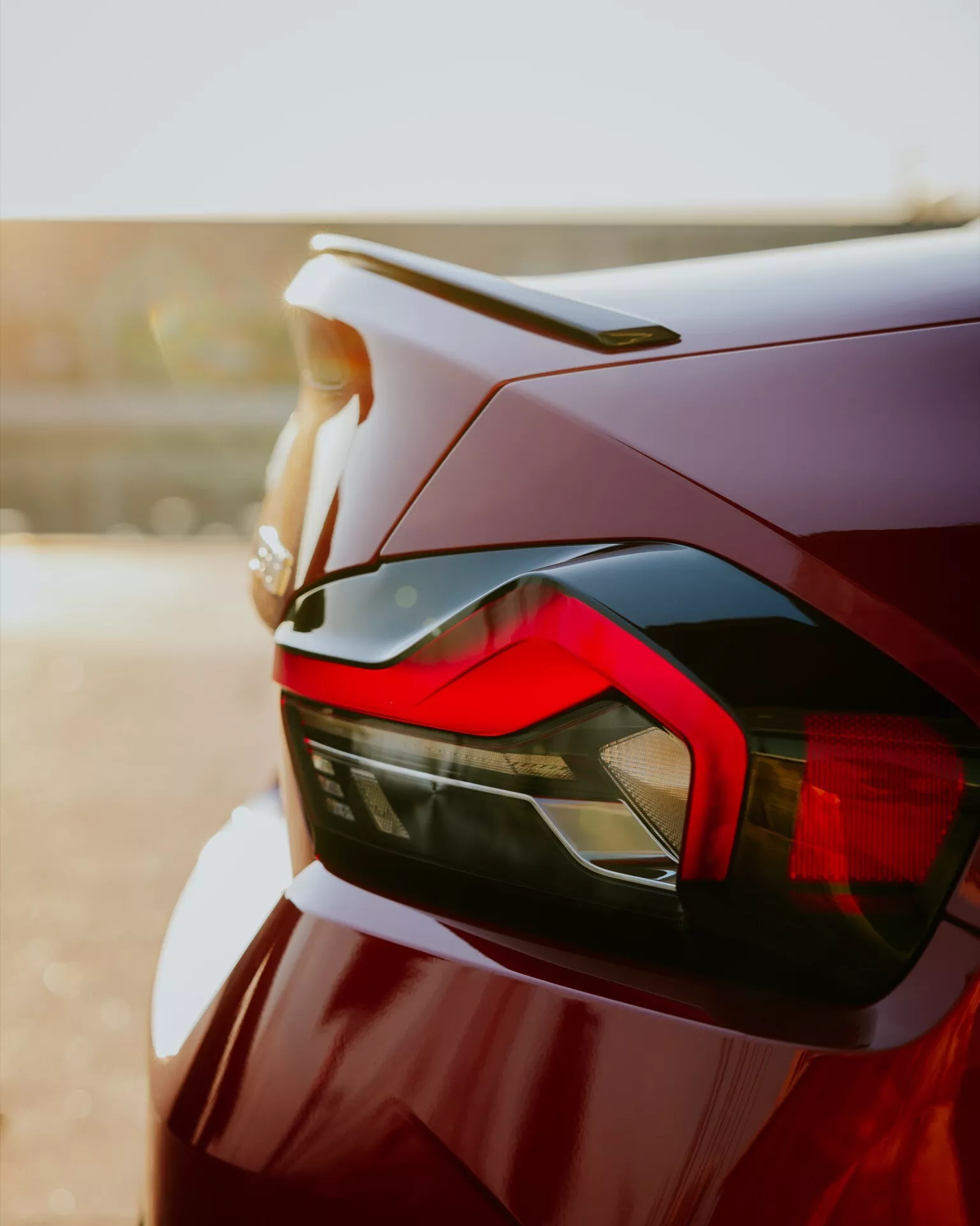 Close-up shot of the rear section of a 2024 BMW 2 Series (G42-G87 facelift), emphasizing the vehicle's design elements such as the sleek taillight with angular LED signatures, a subtle rear spoiler integrated into the trunk lid, and a deep red or maroon high-quality paint finish. The image, taken from a low angle, highlights the car's aerodynamics and modern aesthetics, with a bokeh background creating a warm glow.