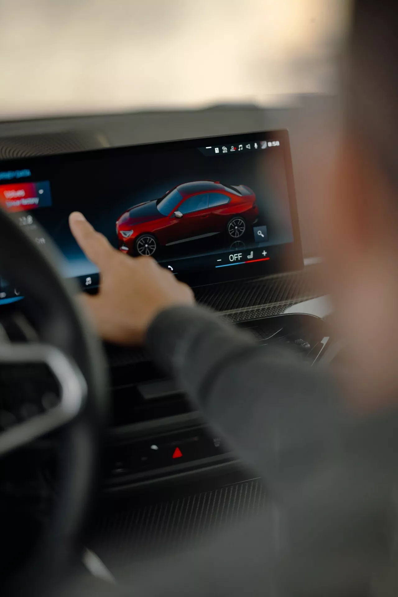 Close-up view of a modern in-car touchscreen infotainment system as seen from the driver's seat, focusing on a customization interface displaying a red sporty car. The steering wheel is slightly blurred, highlighting the high-tech setup of the 2024 BMW 2 Series G42-G87 facelift with advanced features for navigation, settings, connectivity, and multimedia entertainment.