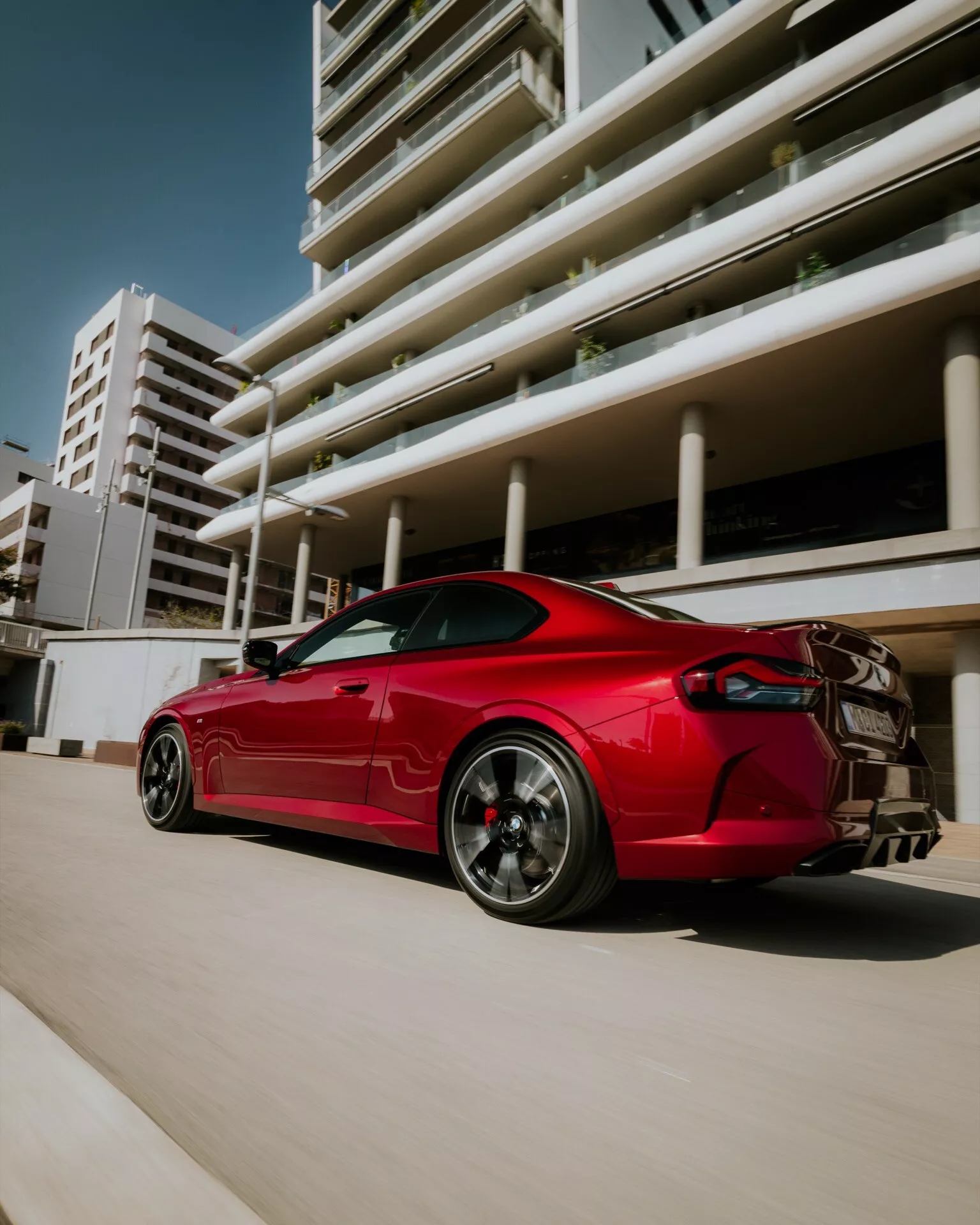 Photograph of the 2024 BMW 2 Series (G42-G87 Facelift) in vibrant red, showcasing a sleek sports coupe from a low rear three-quarter perspective. Features include aerodynamic lines, graphite multi-spoke alloy wheels with visible performance brake calipers, prominent rear spoiler, dual exhaust system, and sharp LED taillights. The image captures the car in an urban setting with modern buildings in the background, emphasizing its aggressive stance and sporty design.