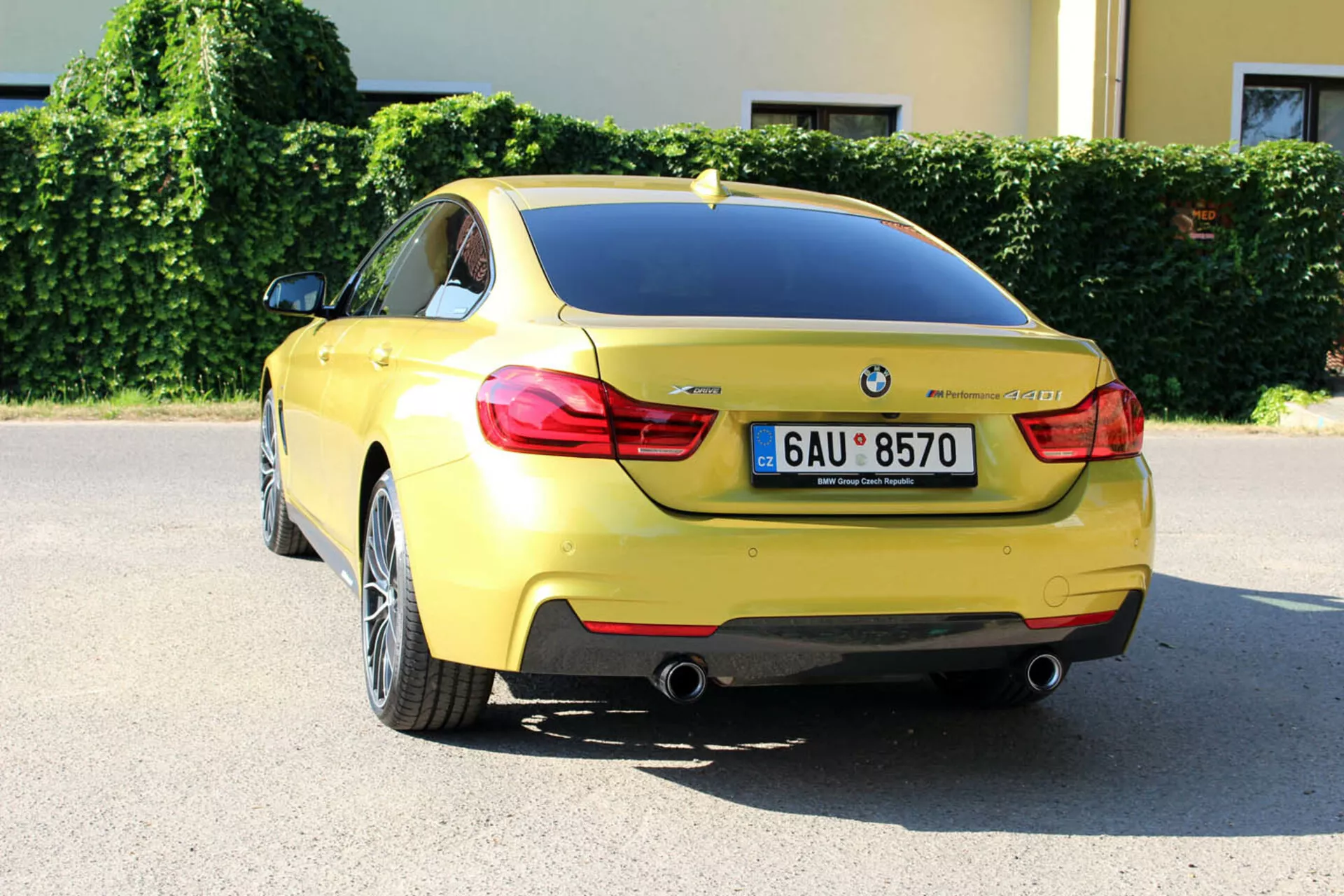 Yellow 2013 BMW 4 Series Coupe from rear three-quarter angle displaying trunk design, rear bumper, partial rear wheel well, distinctive tail lights, dual exhaust pipes, license plate, BMW badge, model designation, parking sensors, shark fin antenna, and multi-spoke alloy wheels, parked in an urban setting on a sunny day.