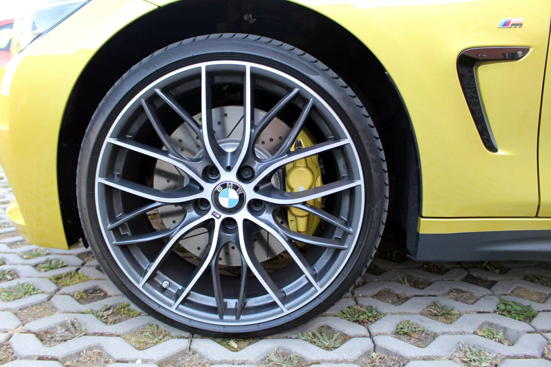 Close-up view of BMW 4 Series Coupe 2013 front wheel featuring multi-spoke dark grey alloy design, low-profile radial tubeless tire, cross-drilled high-performance brake system with yellow calipers, against a bright yellow sporty body