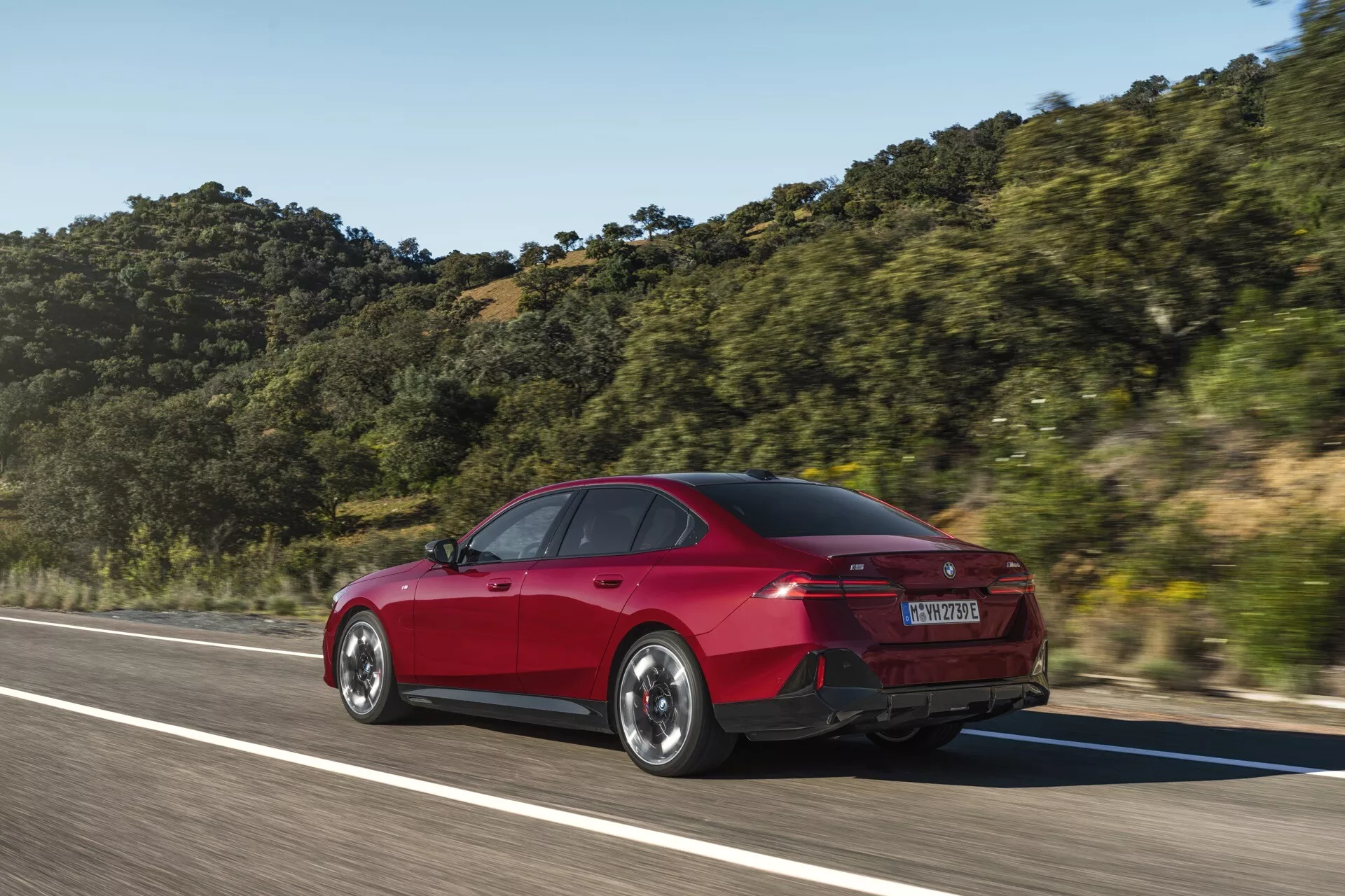 2023 BMW 5 Series (G60-G61) red four-door sedan in motion captured at a three-quarter rear angle, highlighting rear and side design elements like trunk shape, rear bumper, LED taillights, and aerodynamic lines against a mountainous backdrop under clear skies