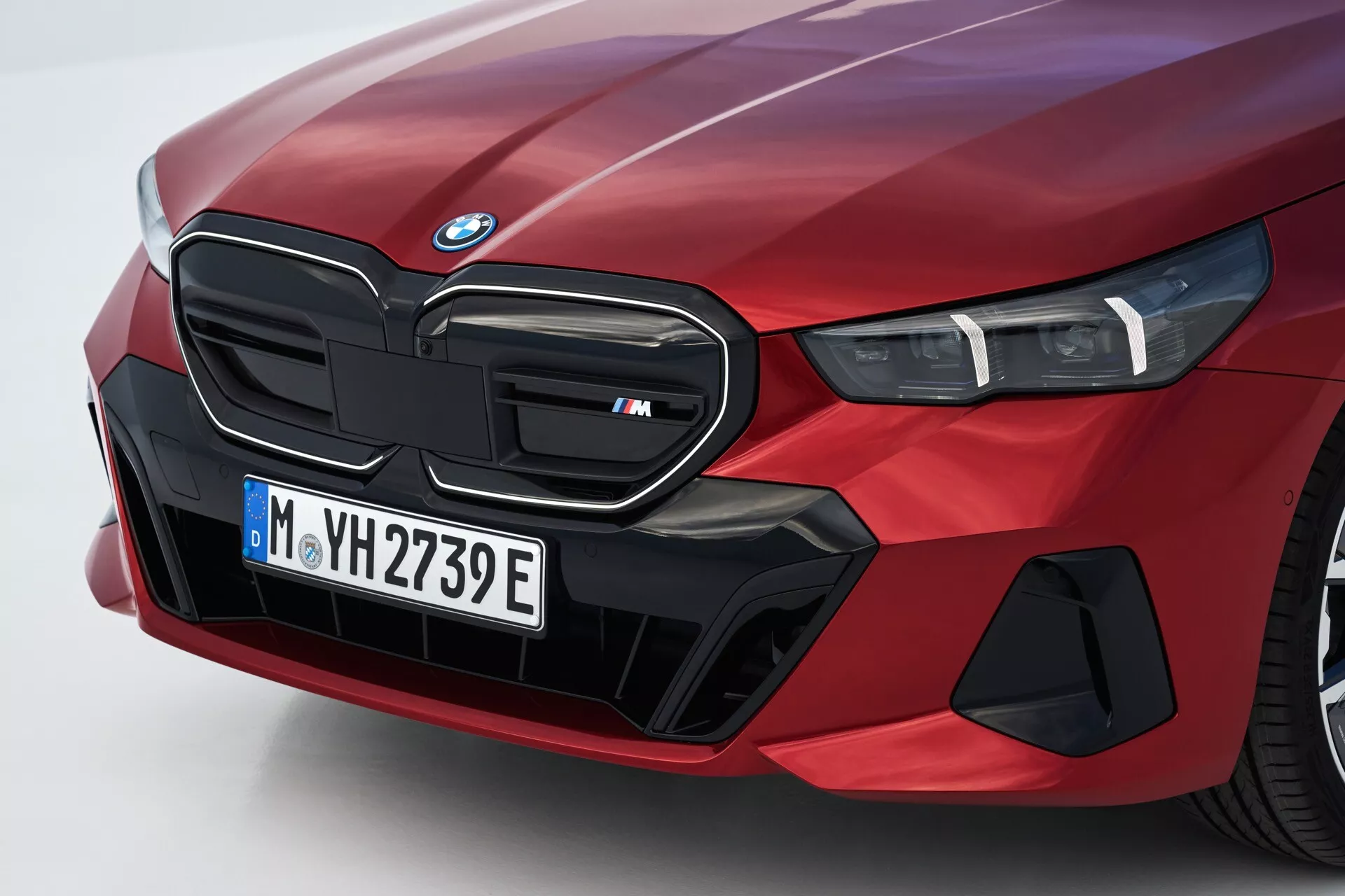 Close-up angled view of the front left side of a 2023 BMW 5 Series (G60-G61) in red, featuring the iconic kidney grille marked as an 'M' model, LED headlight cluster, and specialized aerodynamic elements suggesting electric or hybrid capabilities