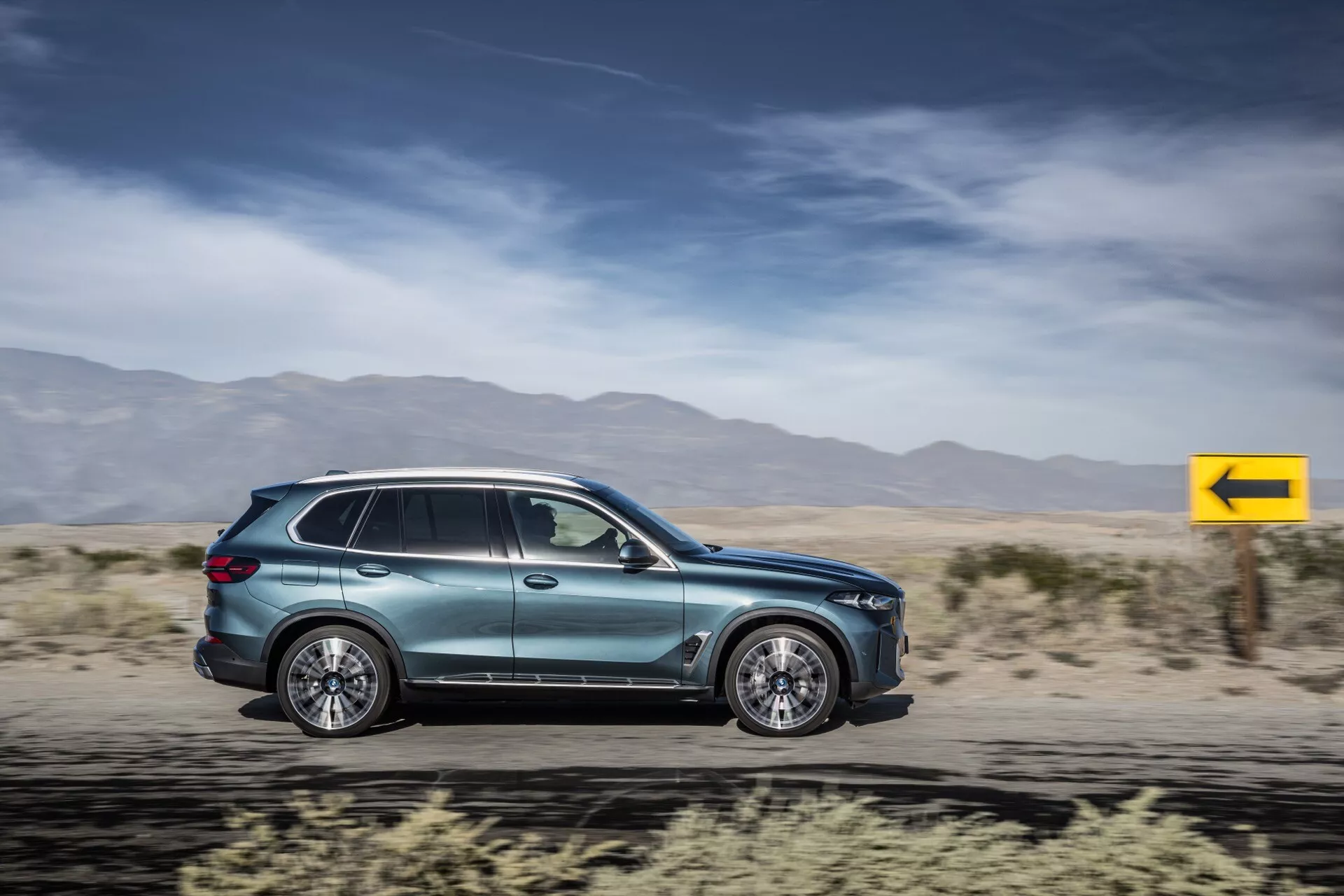 2023 BMW X5 (G05-F95-Facelift) SUV in motion, showcasing aerodynamics, multi-spoke alloy wheels, integrated door handles, aerodynamic side mirrors, tinted windows, side indicator, metallic paint, and modern tail lights design against a motion-blurred desert backdrop.