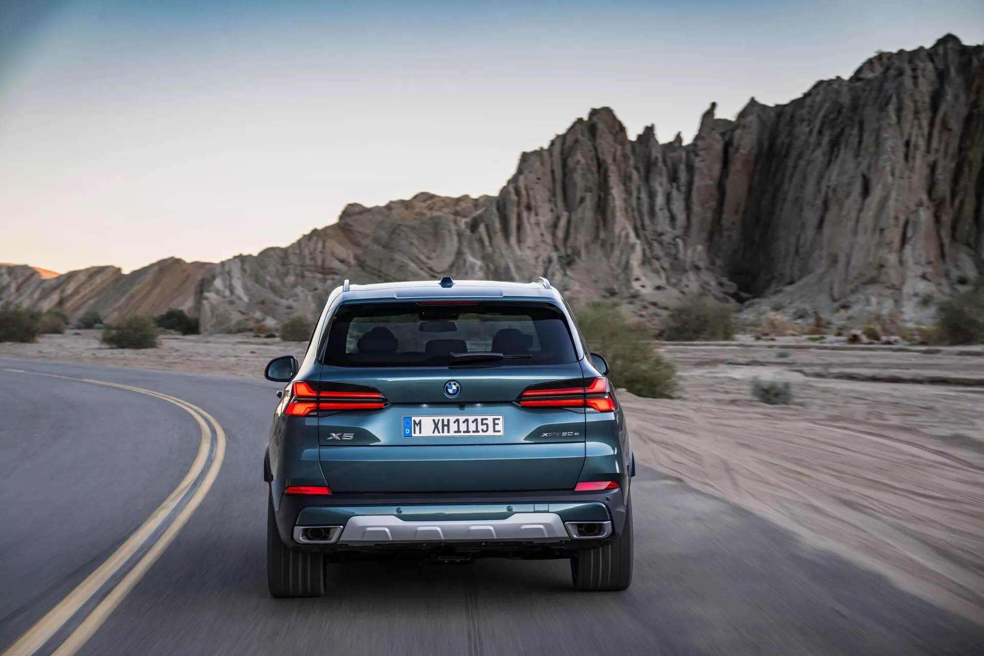 2023 BMW X5 G05 F95 facelift, rear view driving on a curvy road, sporting sleek lines, dual exhaust, and parking sensors set against an adventurous rocky terrain background