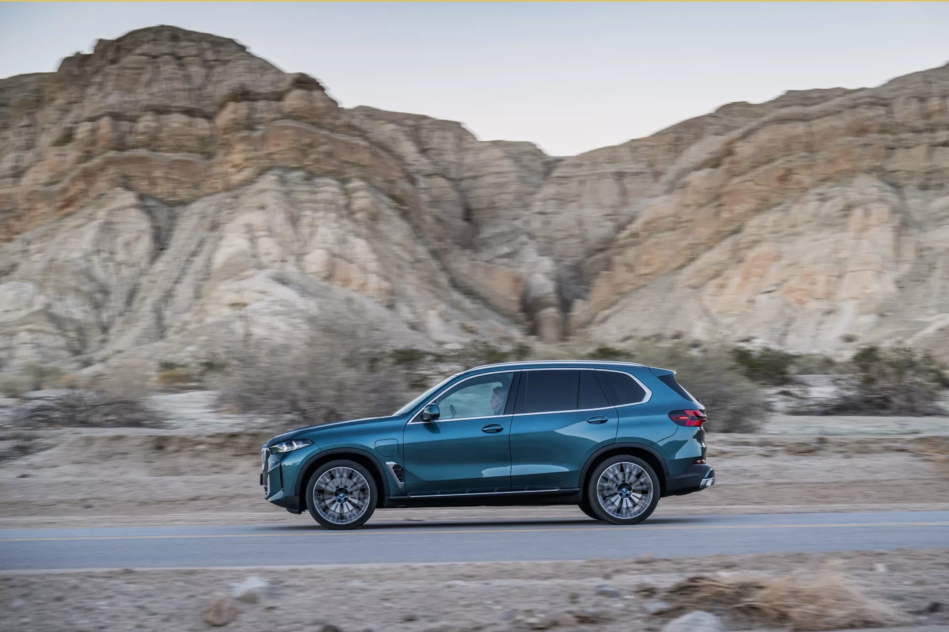 2023 BMW X5 (G05-F95 facelift) dynamic side-profile in motion, blue teardrop-shaped SUV on a road with rugged, barren hills backdrop during golden hour, showcasing sleek design, aerodynamic efficiency, multi-spoke alloy wheels, and LED lighting