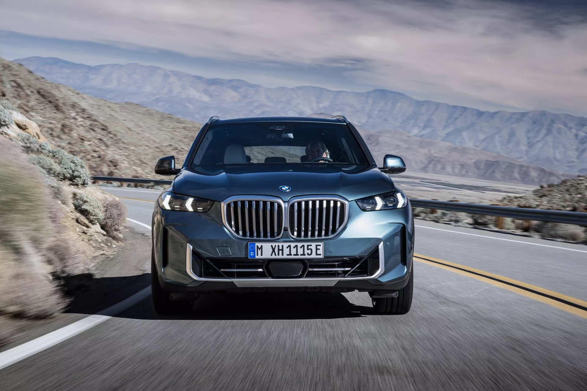 2023 BMW X5 (G05 F95 facelift) in motion, captured at a front three-quarter angle highlighting its LED headlights, kidney grille with active shutters, alloy wheels, and the sleek aerodynamic design against a mountainous desert backdrop, suggesting advanced technology and potential electric or hybrid capabilities