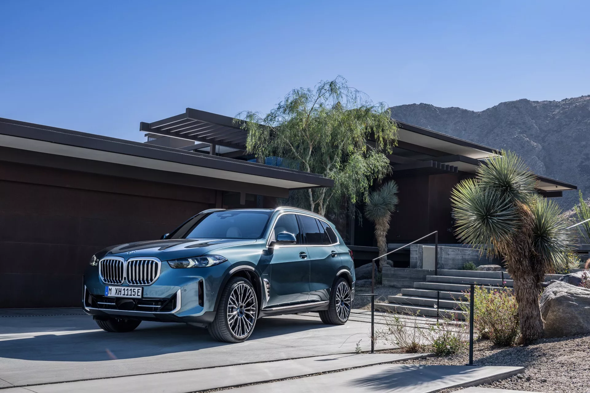 2023 BMW X5 (G05-F95 Facelift) luxury SUV parked in front of a contemporary house with desert and mountain landscape in the background, featuring a metallic finish, large grille, LED headlights, and multi-spoke alloy wheels
