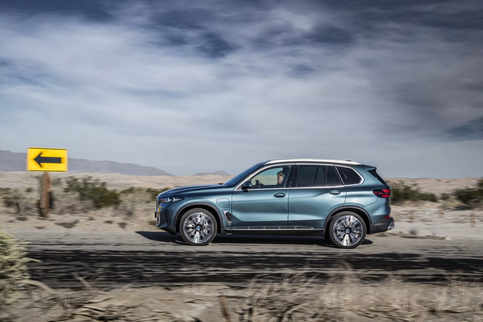 2023 BMW X5 (G05-F95 Facelift) side profile in motion, showcasing its metallic paint job, aerodynamic design, luxury alloy wheels, tinted windows with turn signal mirrors, roof rails, and advanced LED technology, set against a desert backdrop with a dynamic yellow left turn sign.