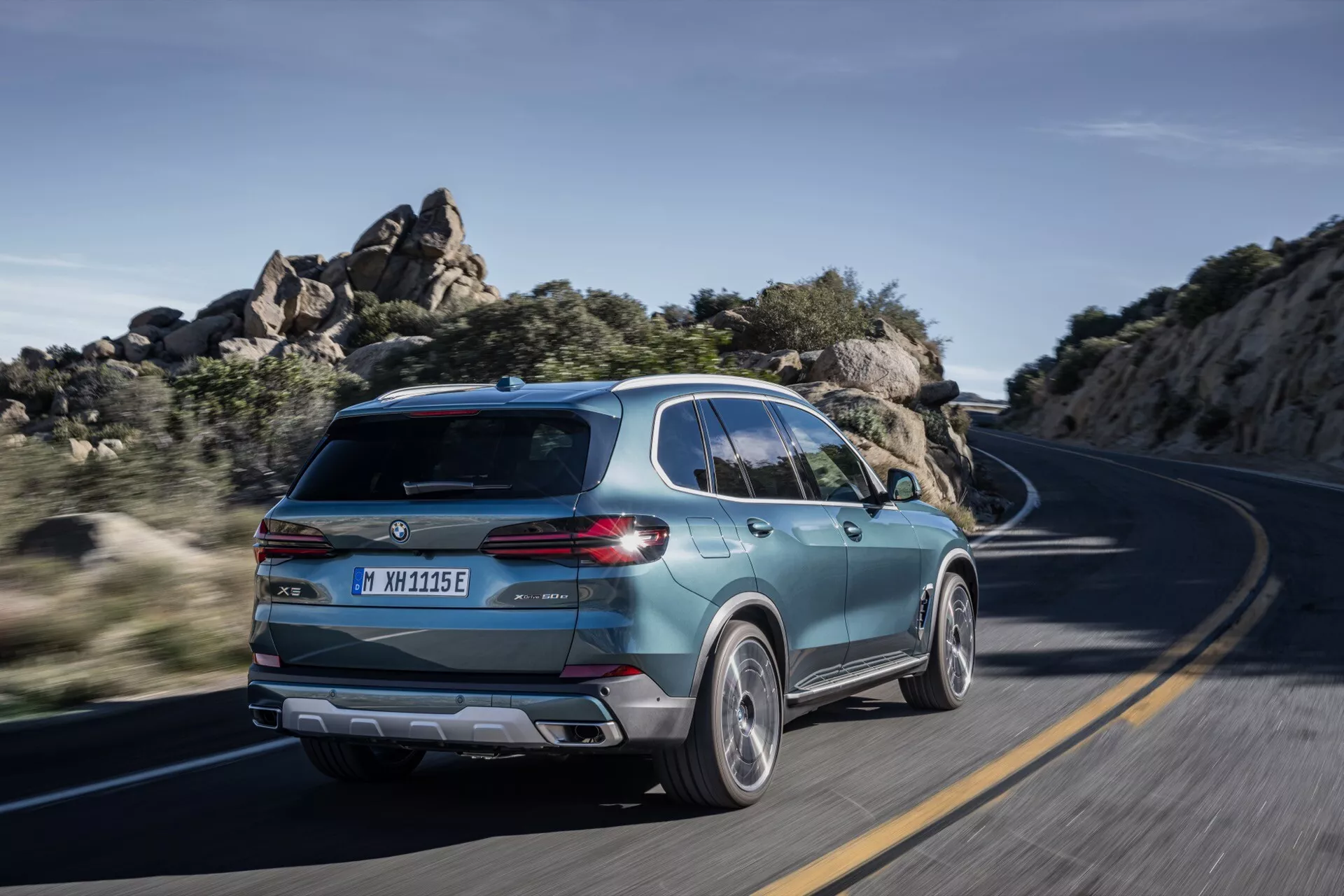 2023 BMW X5 (G05-F95 Facelift) in motion, captured from a rear three-quarter view highlighting its sporty SUV design, modern tailgate with sleek taillights, dual-tone alloy wheels, and evidence of electrification on a winding road under clear daylight
