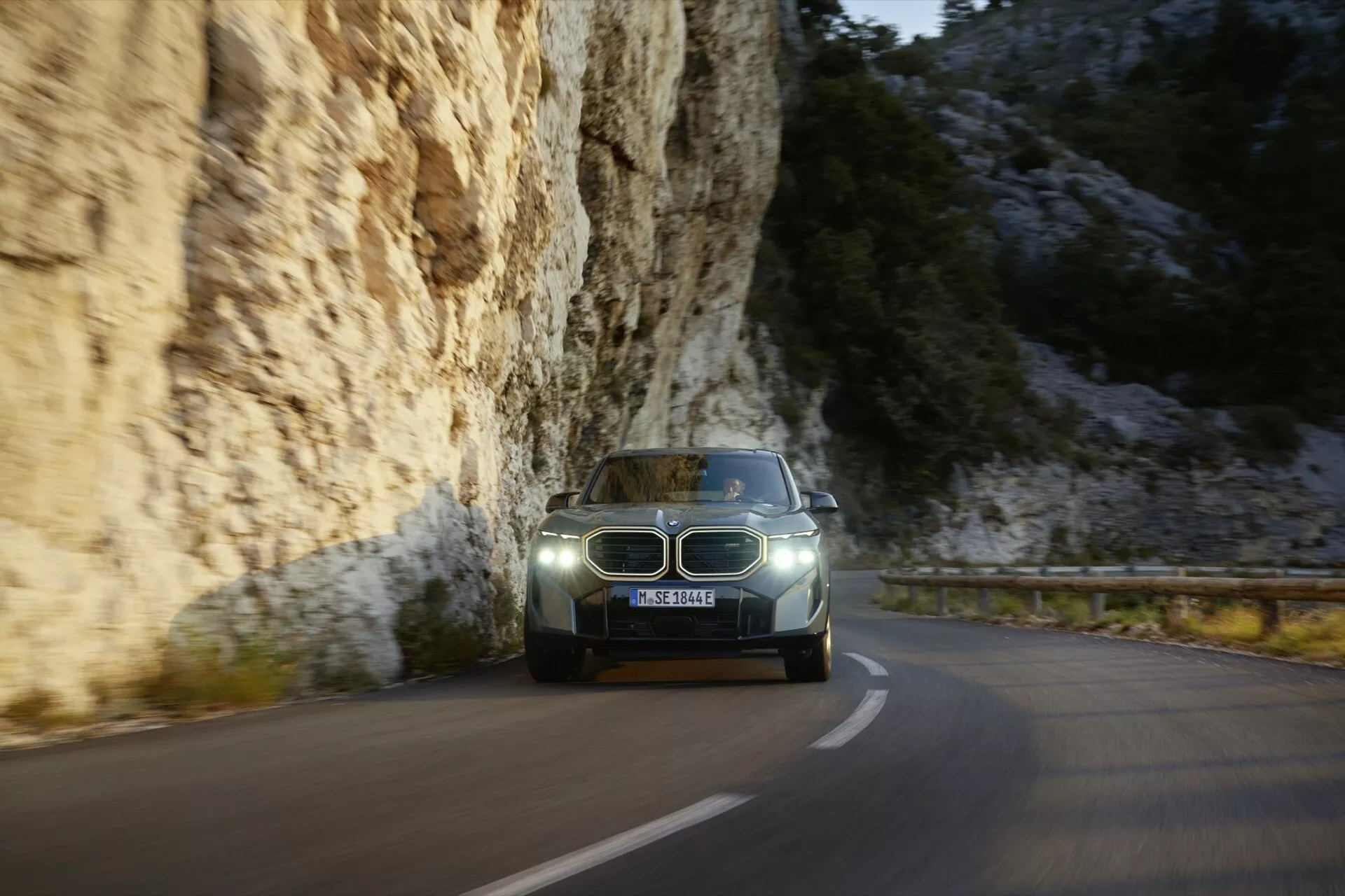2023 BMW XM (G09) driving on a winding mountain road at dusk, featuring LED headlights, illuminated dashboard, and a modern blue-gray paint finish