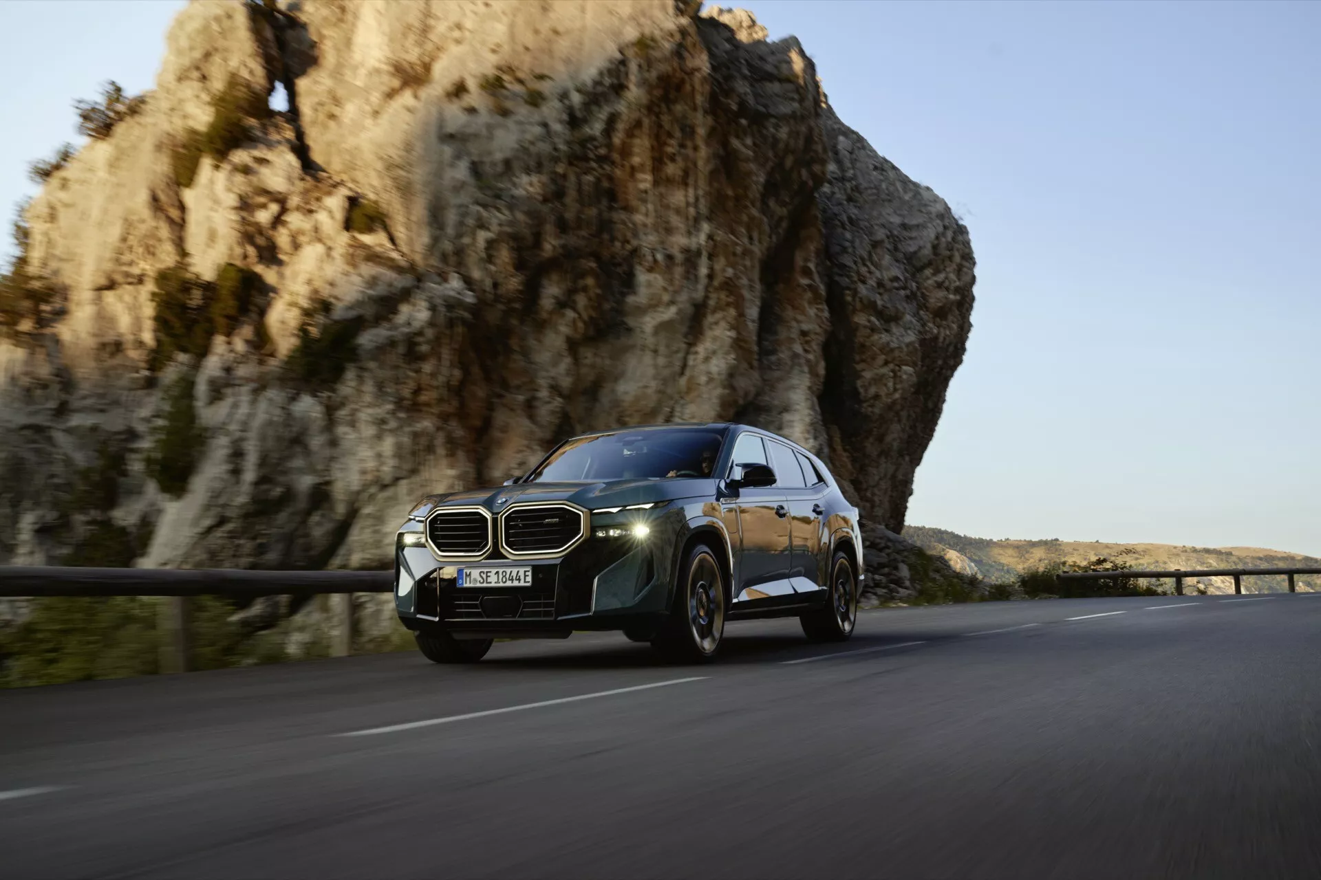 2023 BMW XM (G09) luxury SUV in motion on a scenic route, featuring distinctive kidney grille, modern LED headlights, and aerodynamic design against a backdrop of a rocky cliff and warm sunlight.