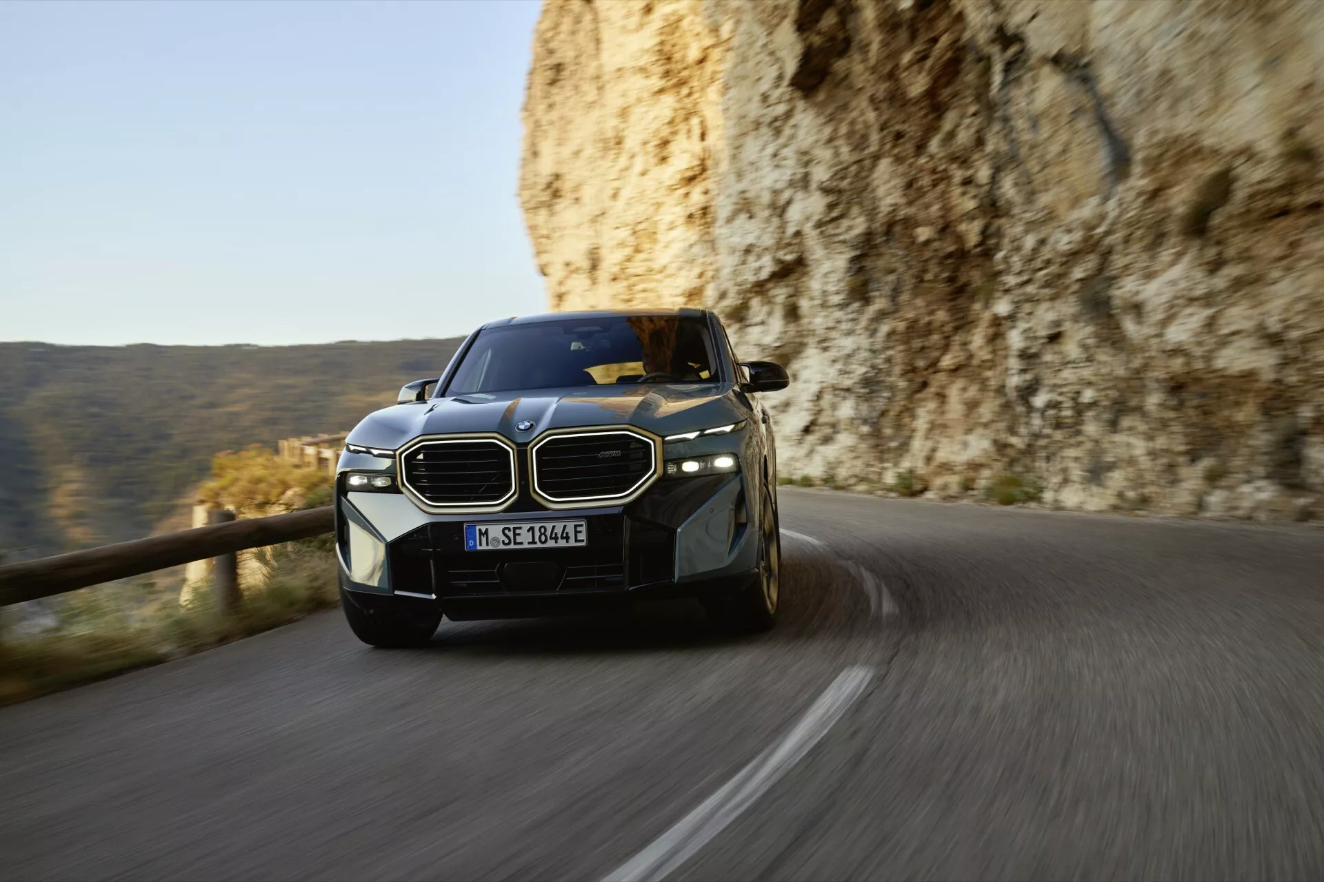 2023 BMW XM (G09) in motion on a mountain road, showcasing its dynamic stance, LED headlights, and distinctive kidney grille from a low front-angle perspective during late afternoon