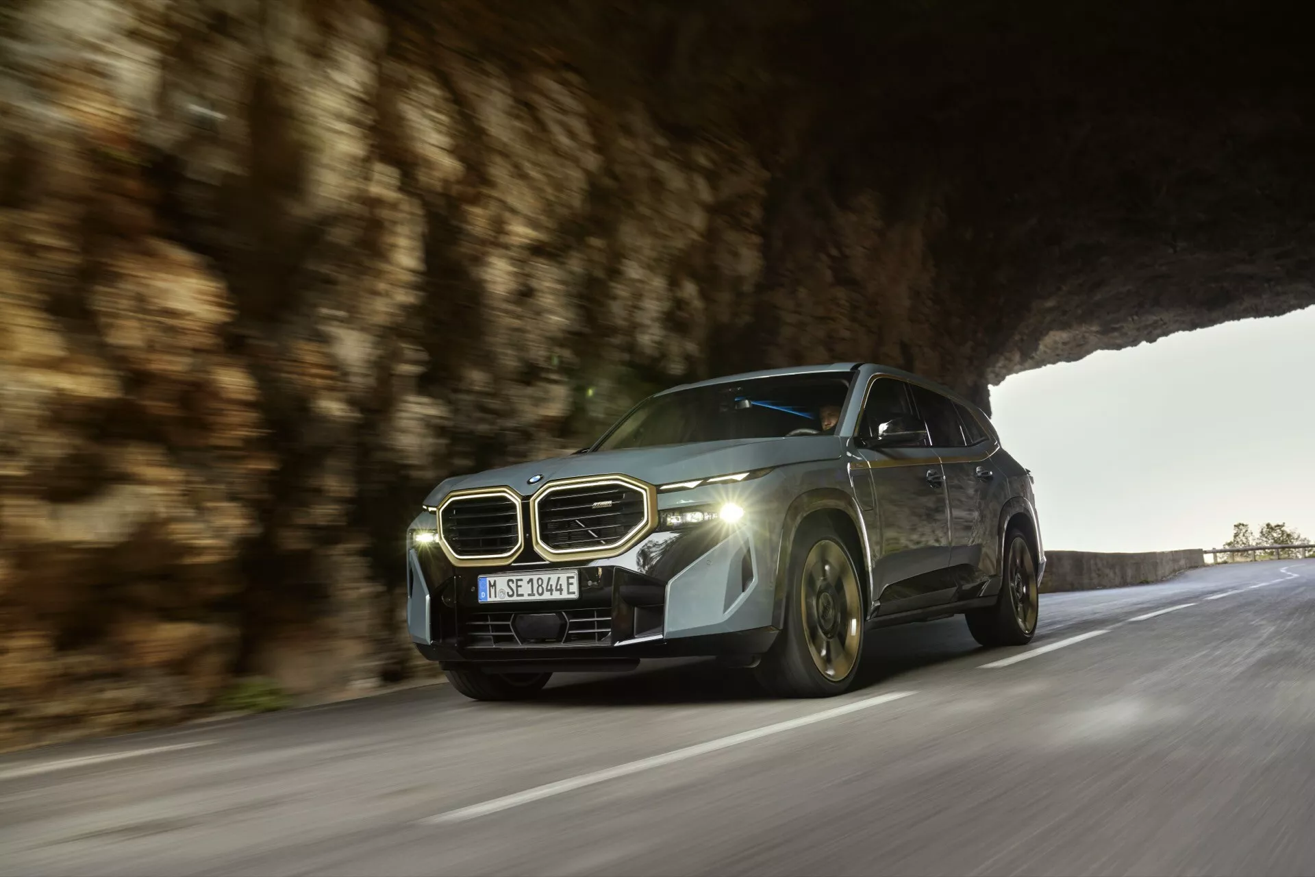 Dynamic low-angle shot of a BMW XM 2023 (G09) in motion on a road, highlighting its modern luxury design, distinctive kidney-shaped grille, emblem, advanced LED headlights, and sleek aerodynamic body with a metallic finish under natural lighting
