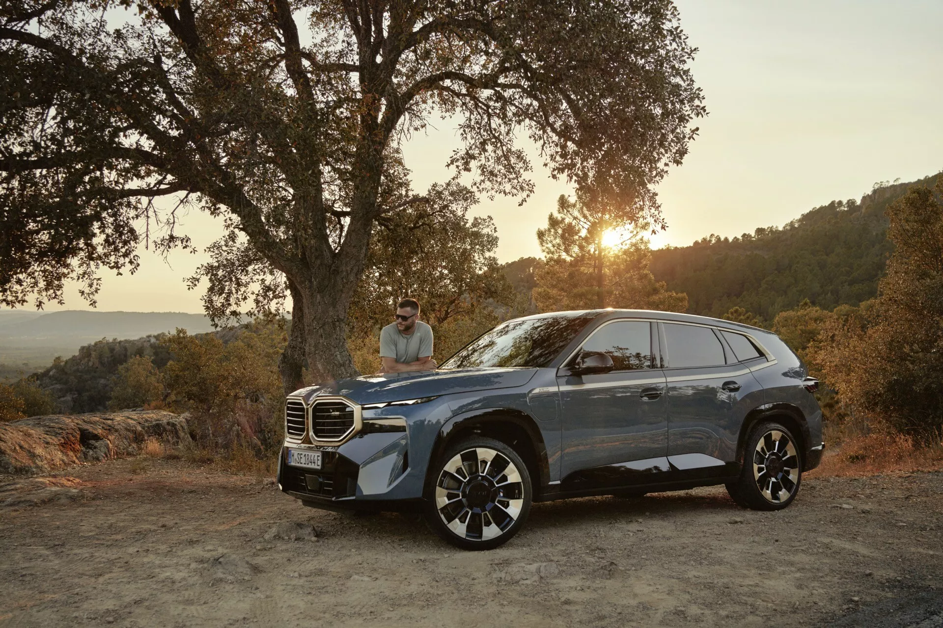 2023 BMW XM (G09) model in metallic finish parked on a dirt road amidst a natural landscape during sunset, showcasing its premium grille, sleek LED headlights, and large alloy wheels from a three-quarter front view