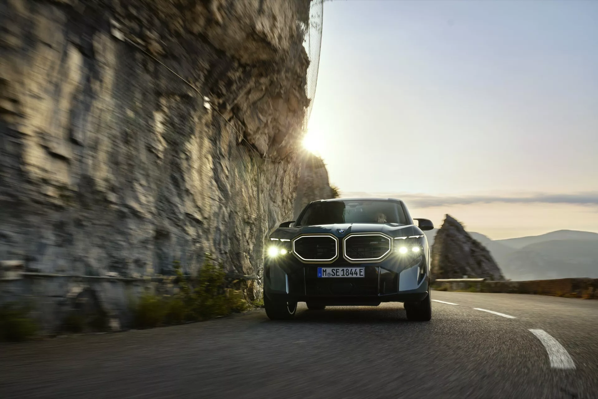 2023 BMW XM (G09) displayed on a winding mountain road at sunrise or sunset, featuring distinctive LED headlamps, iconic kidney grille, potential electric drive system with sensors for driver assistance, and dynamic motion blur indicative of movement.