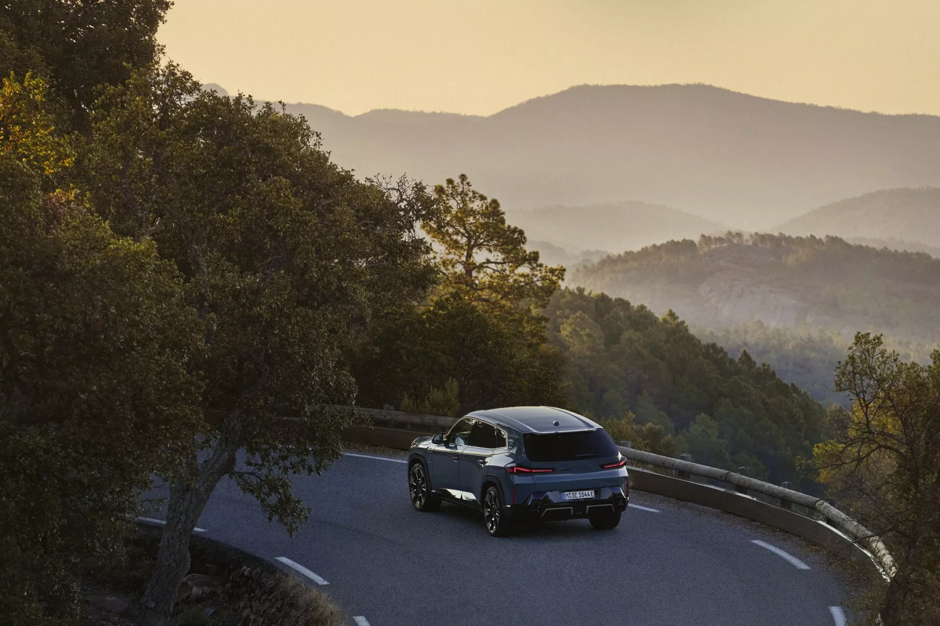 2023 BMW XM (G09) seen from a rear three-quarter angle on a mountainous road during sunrise or sunset, showcasing its sleek design, LED taillights, and alloy wheels