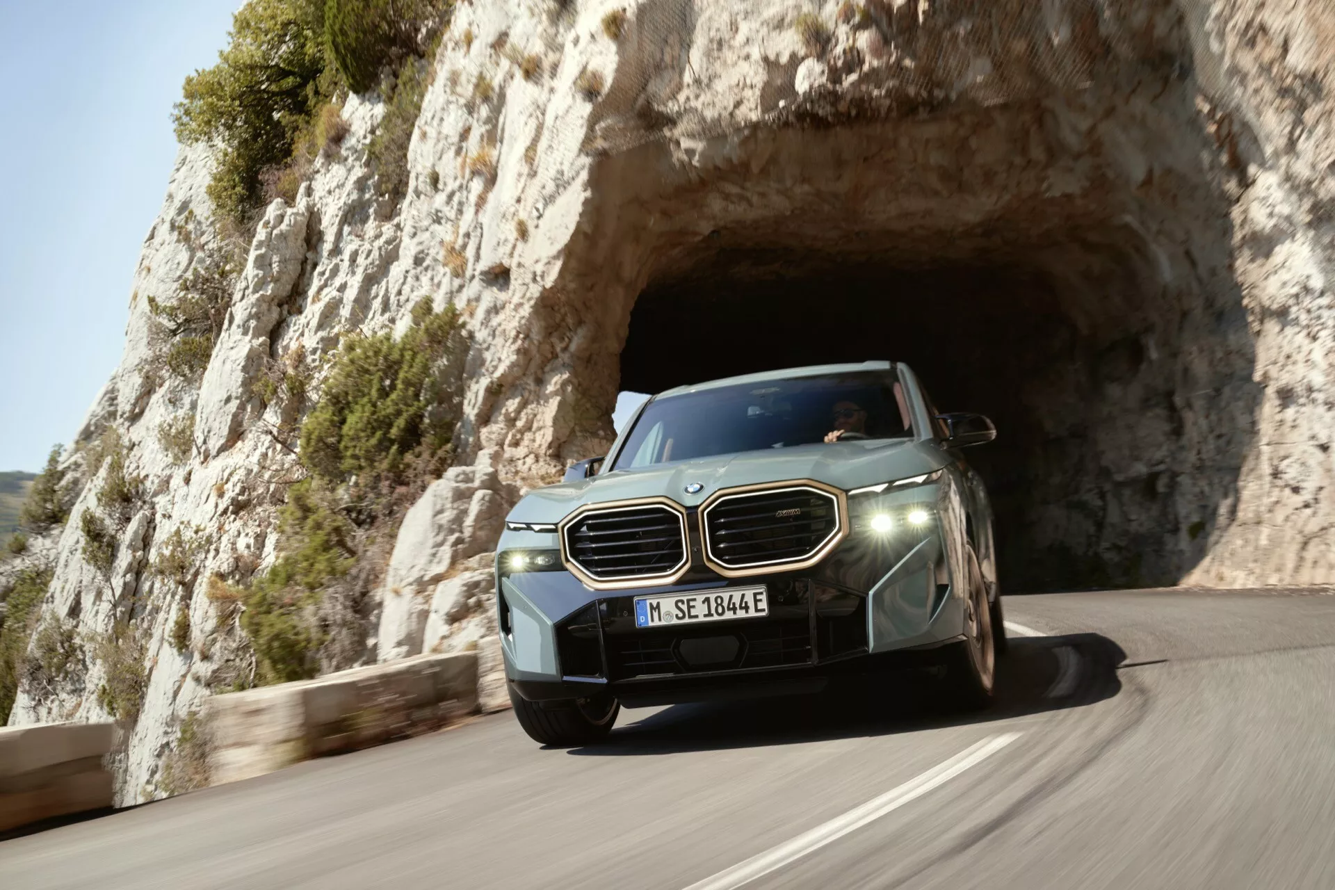 2023 luxury SUV captured in motion exiting a natural rock arch or tunnel, showcasing its aerodynamic design and advanced technological features like LED headlights and driver assistance sensors, with a European license plate, against a scenic coastal or mountainous backdrop.
