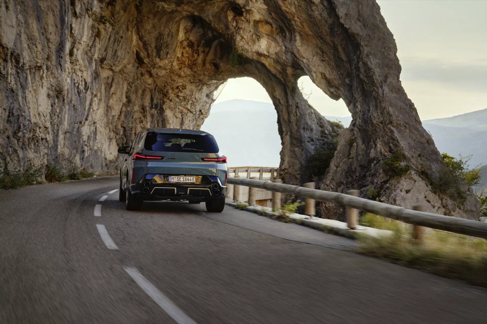2023 BMW XM (G09) captured from a rear three-quarter angle on a mountainous road, highlighting its dual exhaust system, sleek tail lights, rear diffuser for aerodynamics, elevated SUV design, large alloy wheels, rear spoiler, tinted windows, and sporty side profile in an evening setting